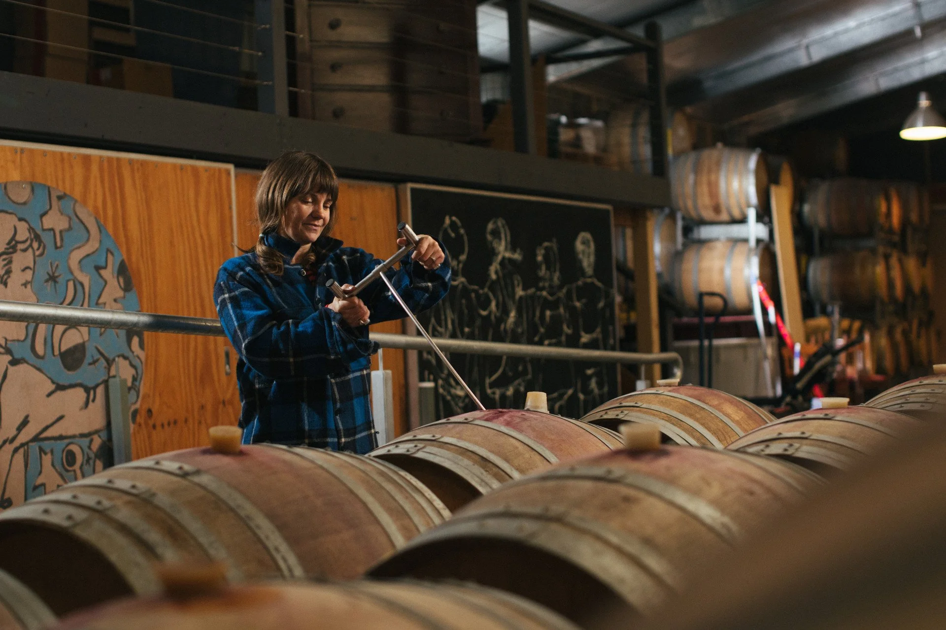 A woman in a blue plaid shirt opens a wine barrel in a wine cellar with wooden barrels and a blackboard in the background.