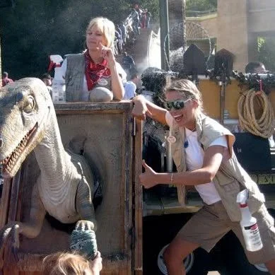 Two smiling white women at Universal Studios California in safari costumes. One stands behind a life-sized velociraptor puppet wile the other stands next to it wearing sunglasses and giving an enthusiastic thumbs up.