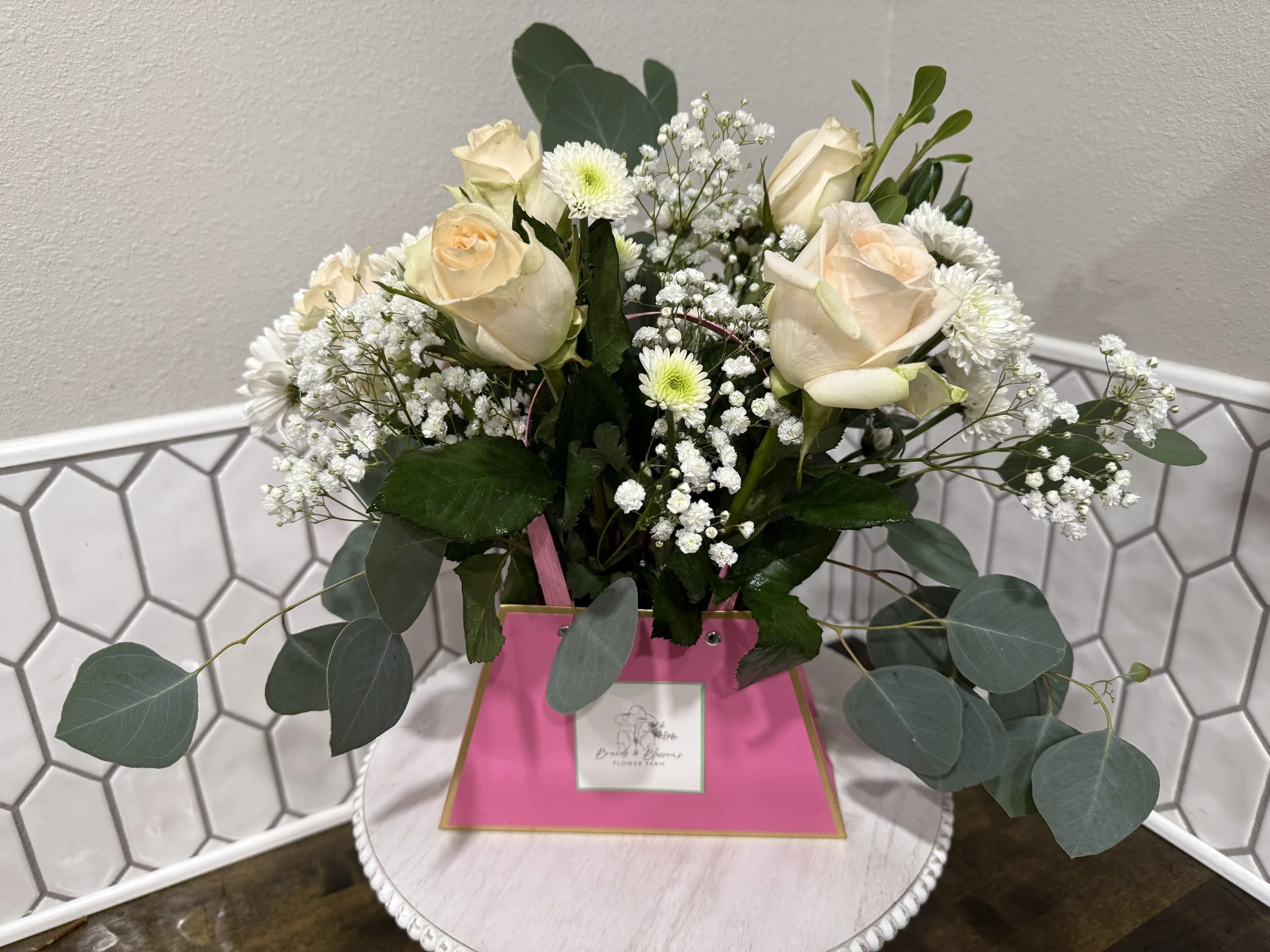 A floral arrangement with white roses, baby's breath, and white chrysanthemums in a pink flower box on a white round table.