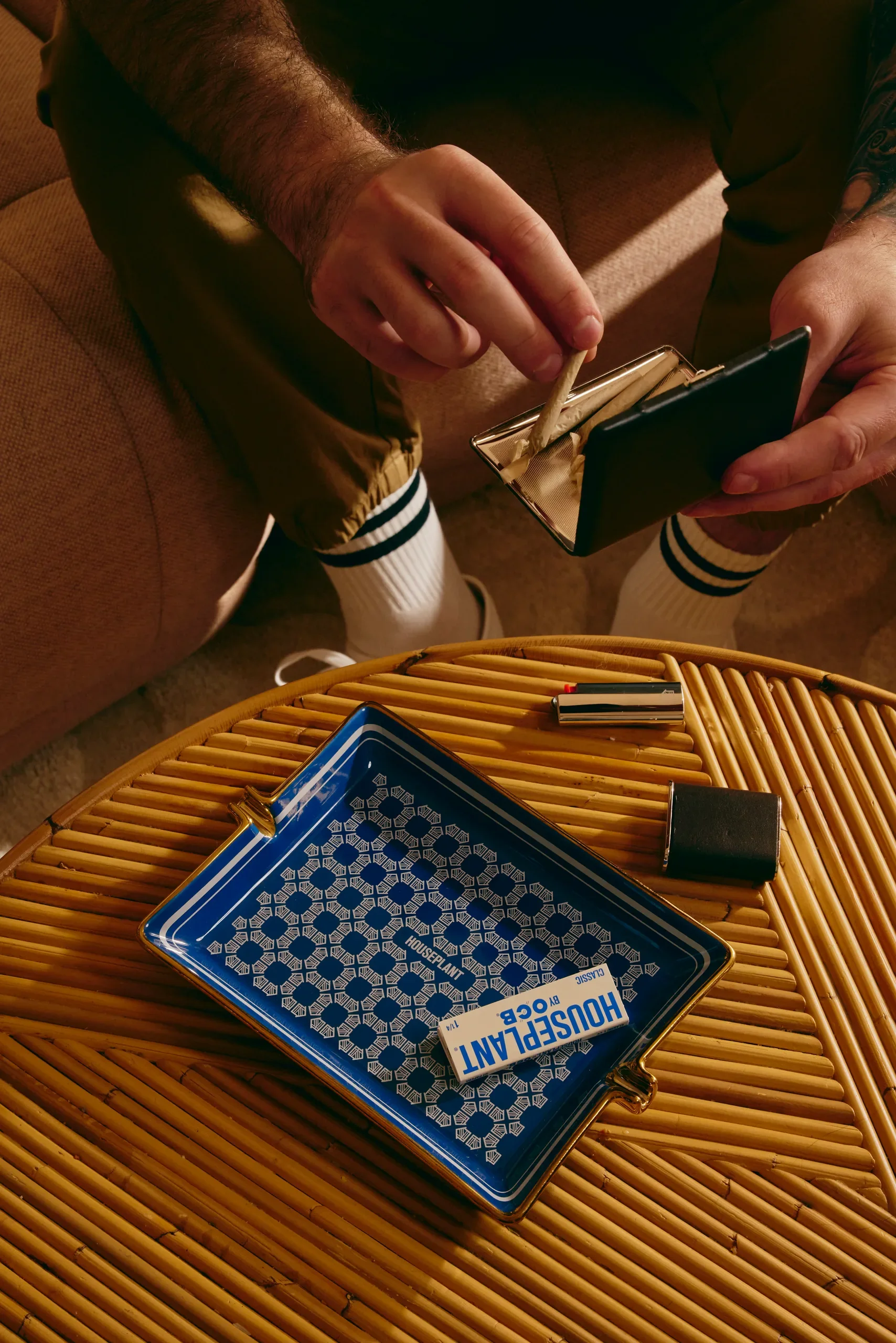 Person pulling a pre-rolled joint from a black metal case over a wicker table with a blue Houseplant by OCB ashtray.