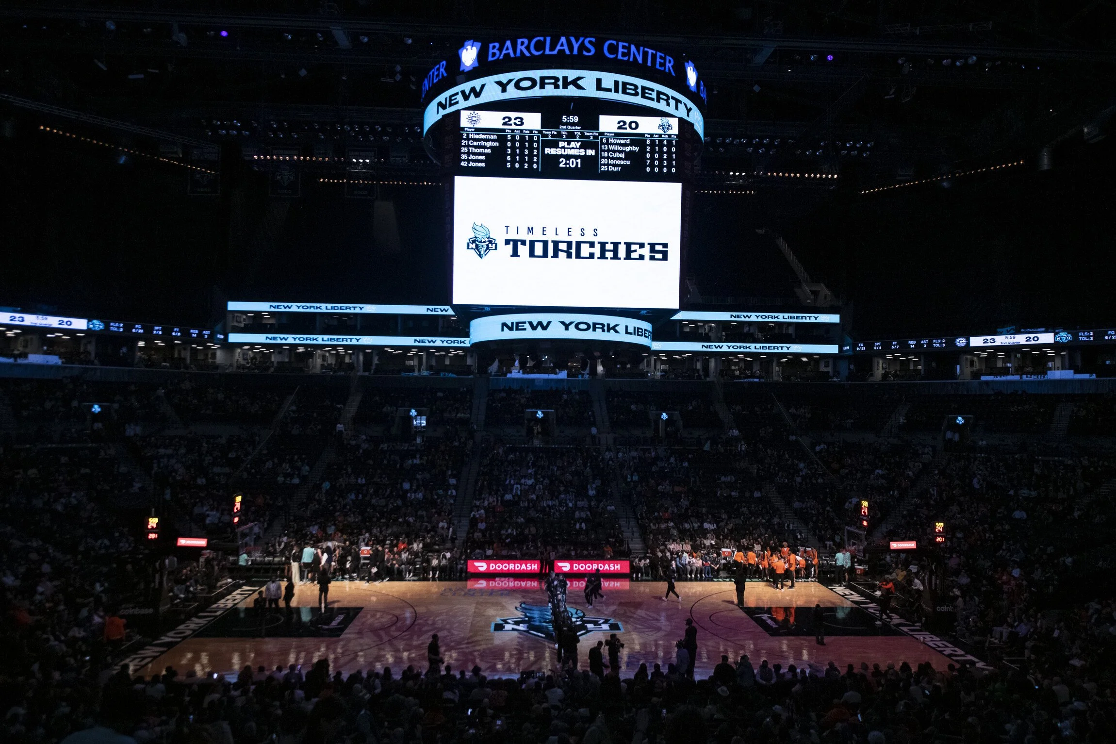 Inside of a basketball arena with the scoreboard showing the New York Liberty versus the opponent, and the text "Timeless Torches" on a large screen. The game is ongoing, with the score 23-20 and 5:59 remaining in the second quarter.