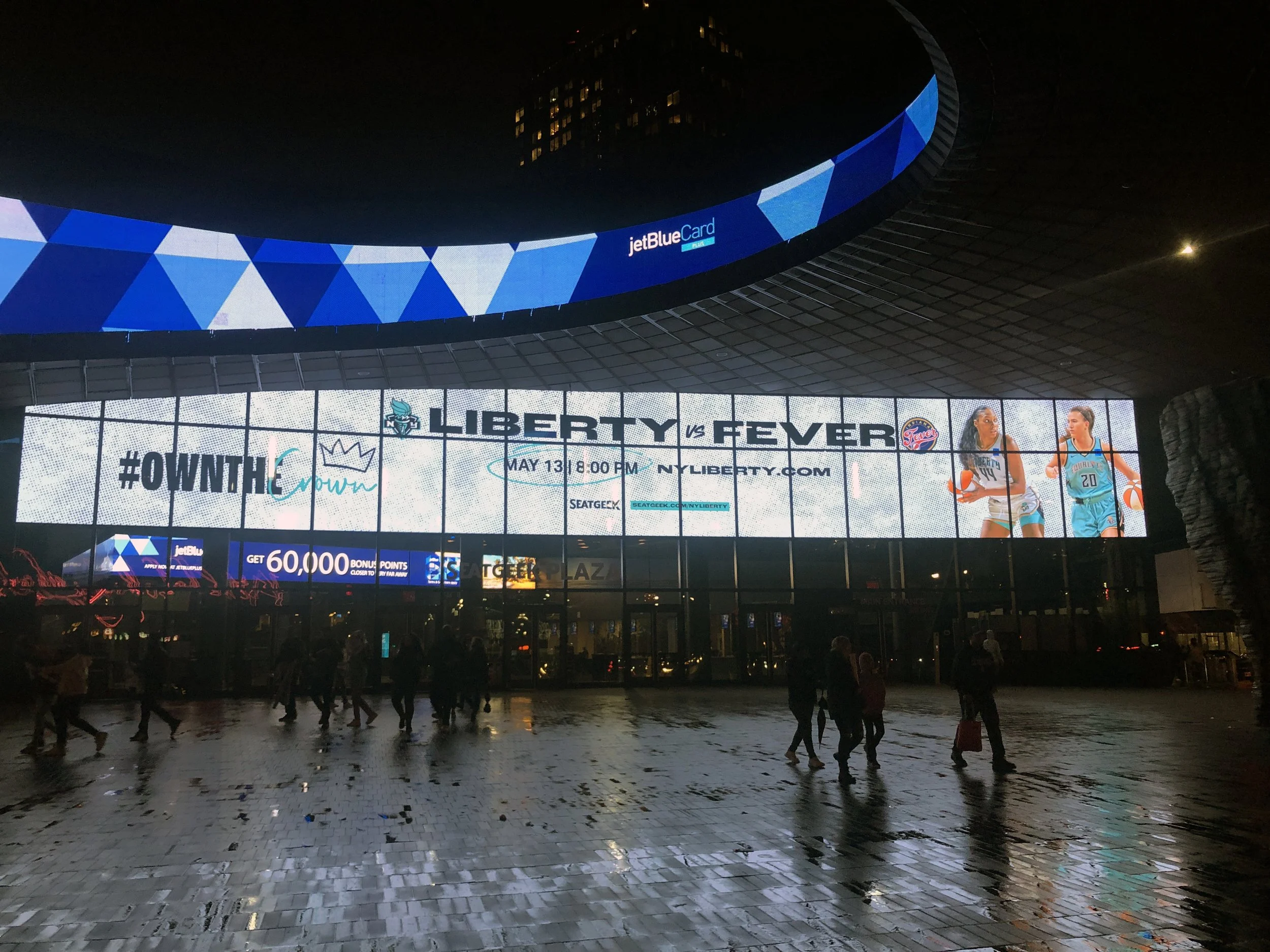 Digital billboard advertising a basketball game between Liberty and Fever on May 13 at 8:00 PM, with images of female basketball players, located outside near a wet pavement at night.