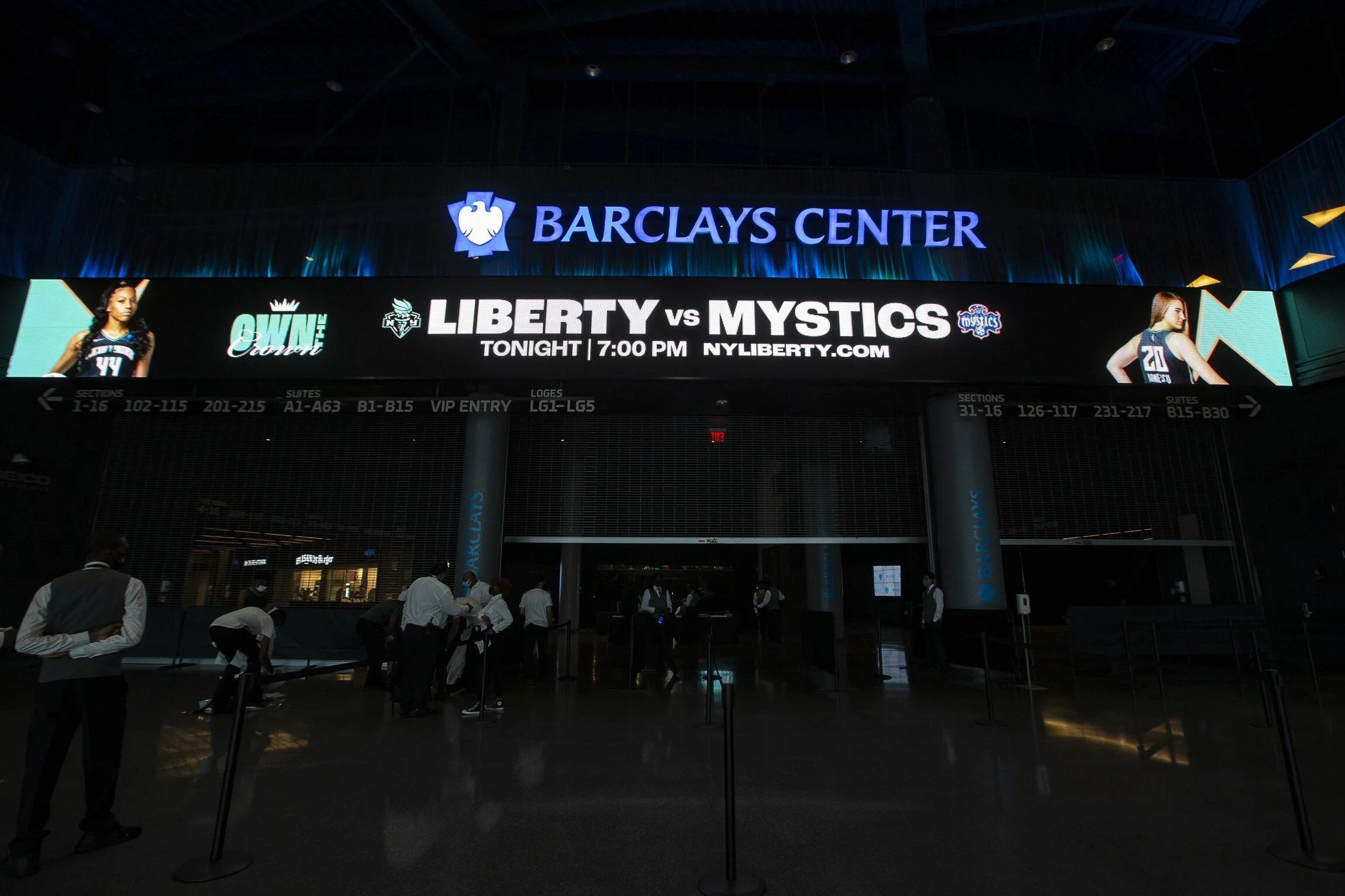 The entrance area of Barclays Center with a digital promotional banner for a Liberty basketball game against Mystics featuring two female players, and several people waiting in line.