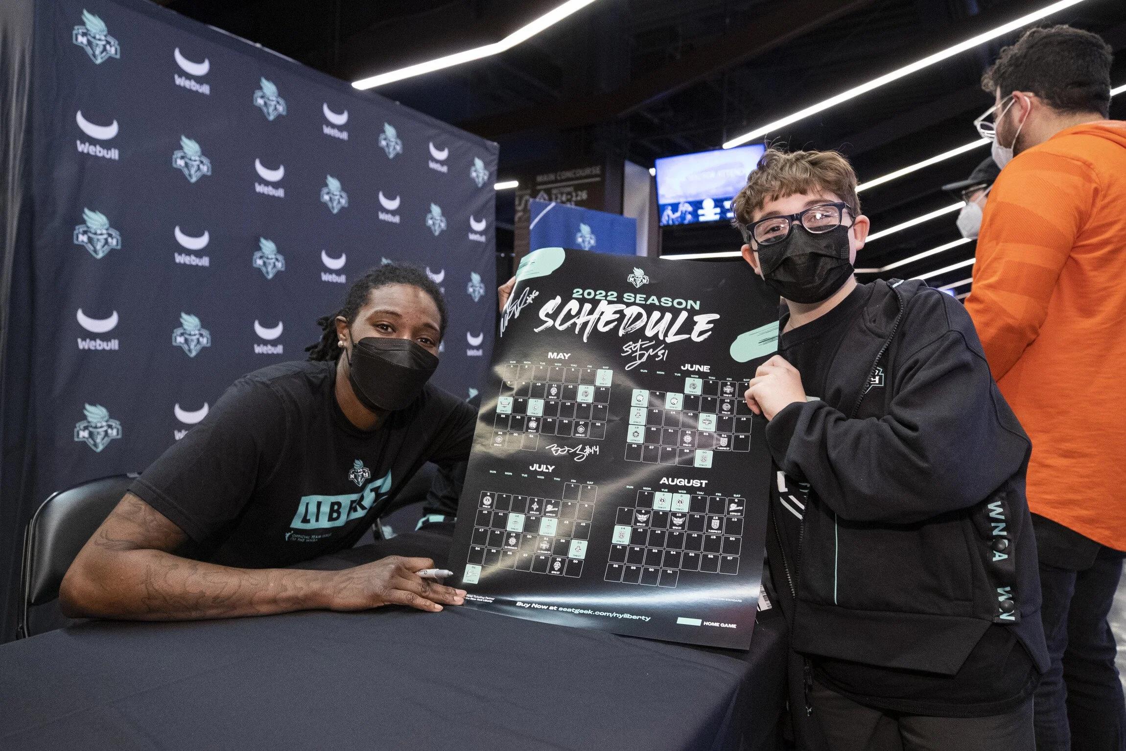 Two people wearing masks holding a 2022 season schedule poster at an event, with a backdrop featuring the Webull logo and a person in an orange jacket in the background.