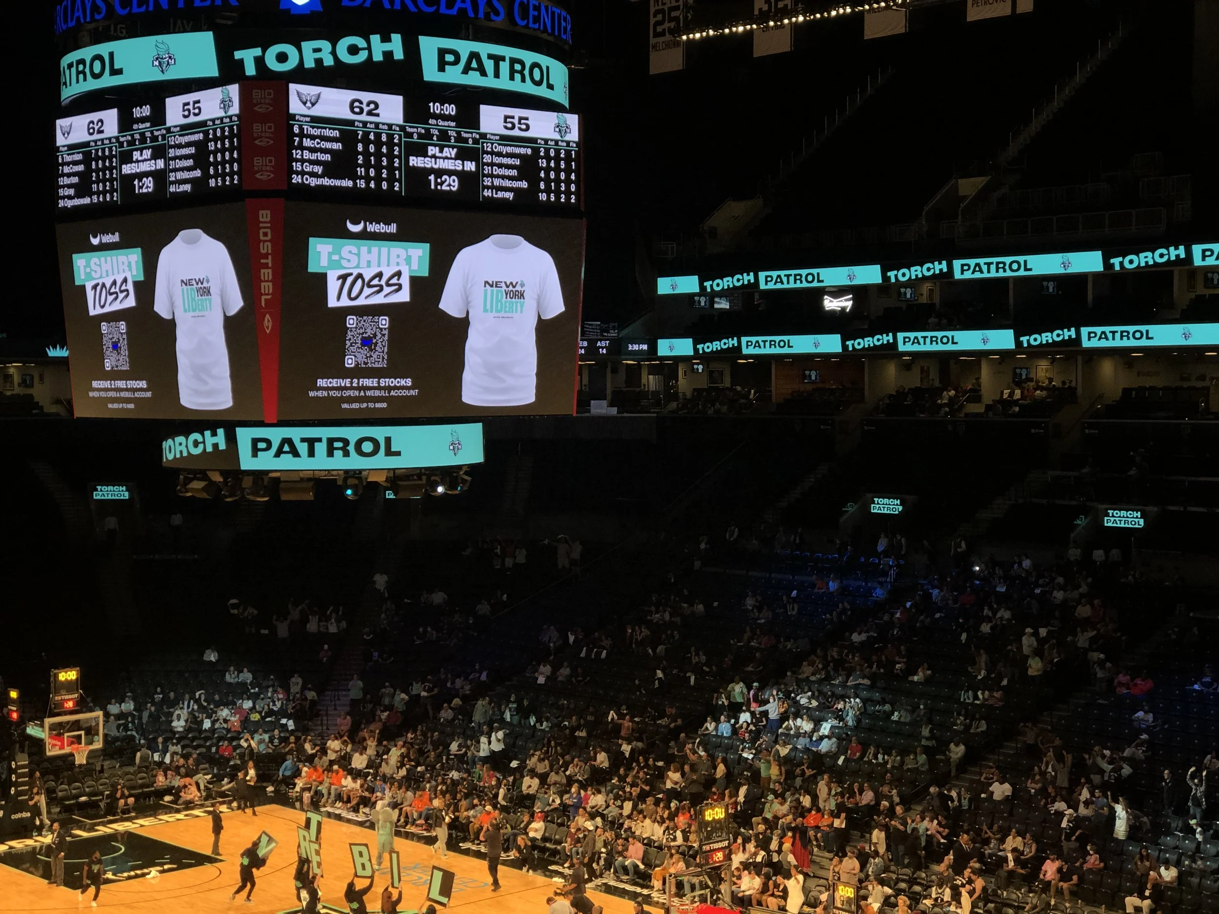 Indoor basketball arena with audience seated in the stands. Large digital scoreboard displaying the current game score (62-55) with 1 minute and 29 seconds remaining. Multiple digital banners and screens showcase advertisements for free T-shirts and 