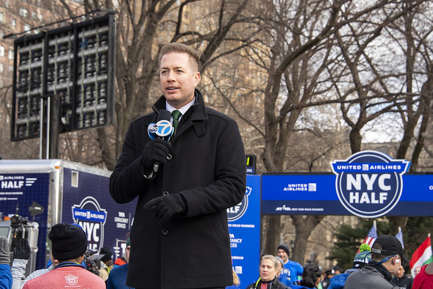 A man in a black coat and gloves speaking into a microphone with the ABC 7 logo at an outdoor event. Behind him is a blue sign that reads 'NYC Half' and 'United Airlines' with many people, some wearing winter clothing, gathered at what appears to be 