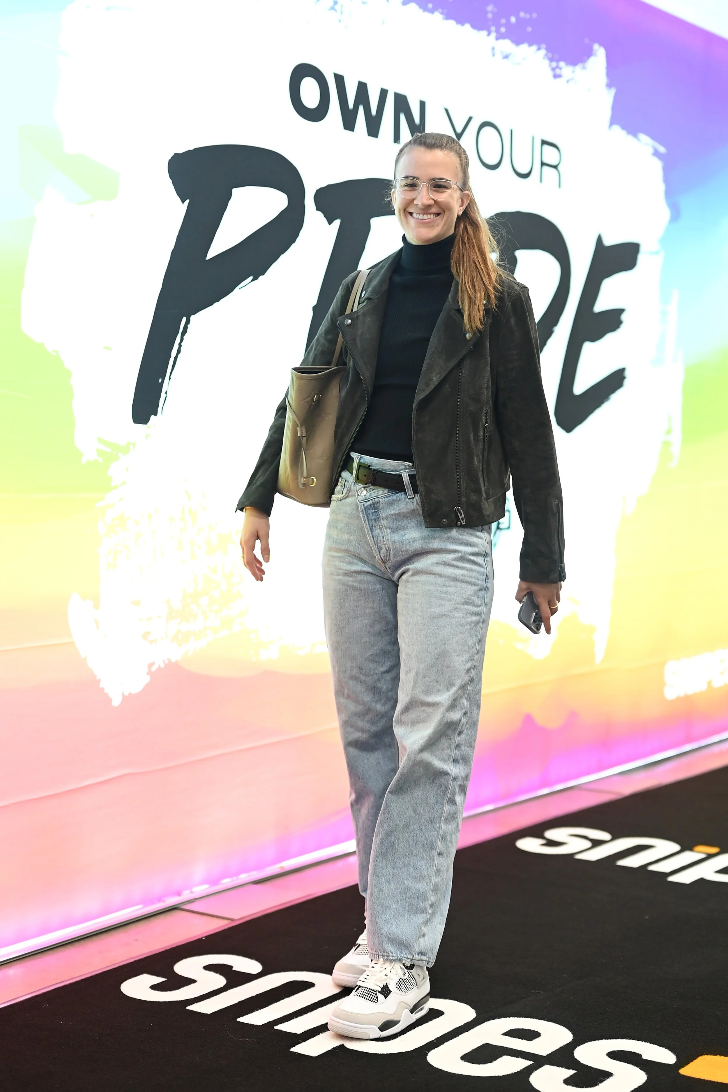 Smiling woman in black turtleneck, black leather jacket, light wash jeans, and white sneakers standing in front of a colorful backdrop with the words 'Own Your Power.'