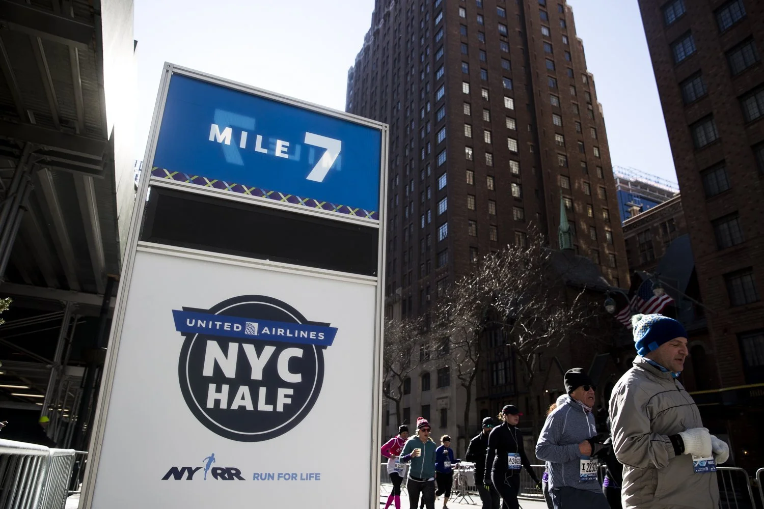 Runners participating in a half marathon in New York City, with a sign indicating mile 7 and the event's name, NYRR NYC Half, sponsored by United Airlines.