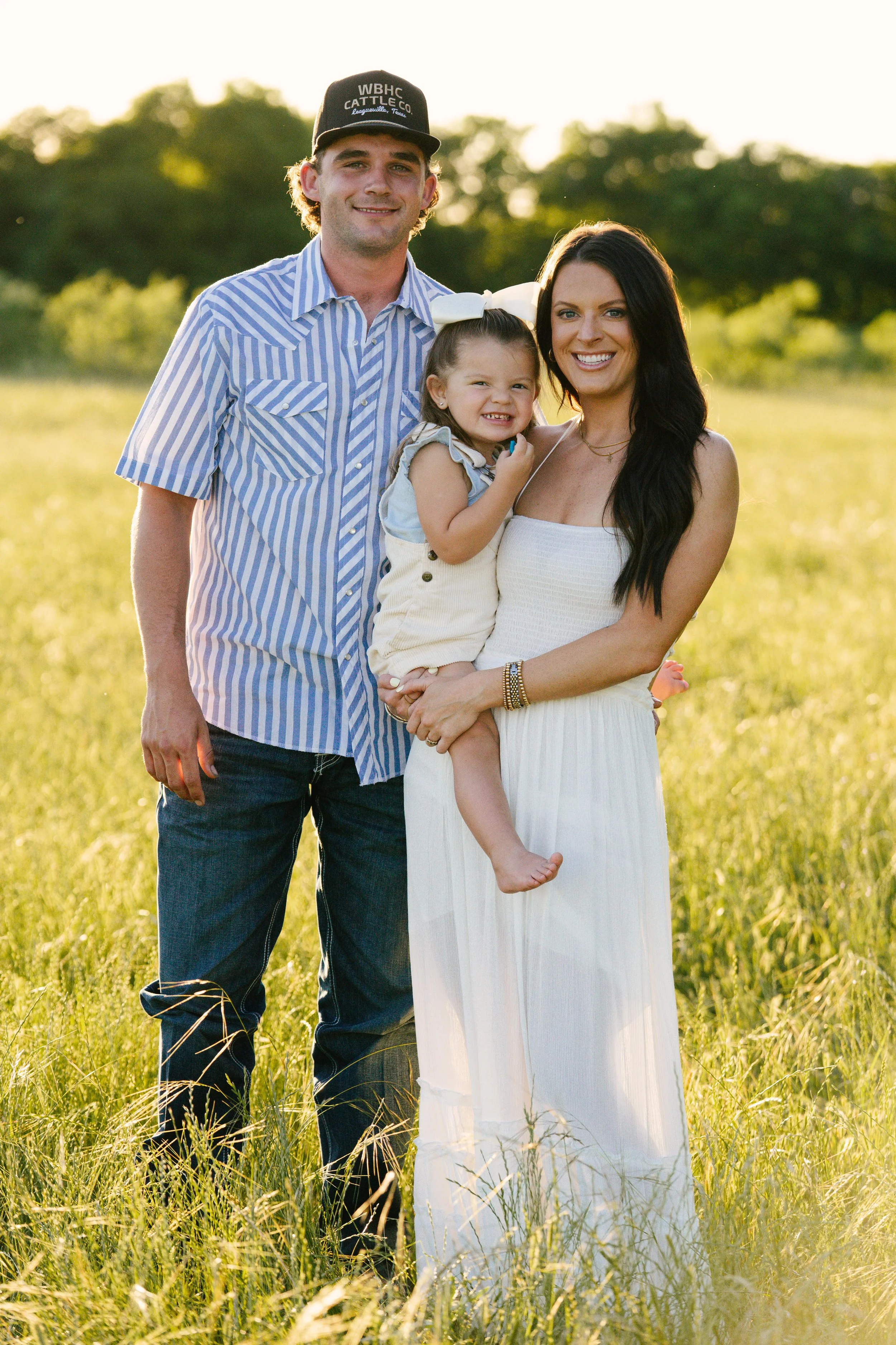A family of three standing outdoors in a grassy field during sunset, smiling at the camera. The father is wearing a striped shirt and a black cap, the mother in a white dress holding their young daughter, who has a large white bow in her hair.