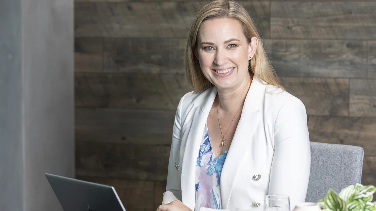 A woman with blonde hair smiling and sitting at a table with a laptop.