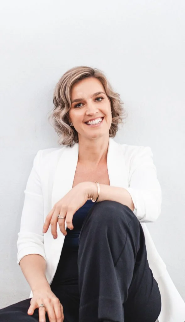 A woman with short, wavy blond hair, smiling, sitting against a white wall, wearing a white blazer and black pants.
