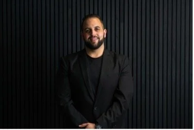 A smiling man with a beard in a black blazer standing in front of a dark, vertically striped background.
