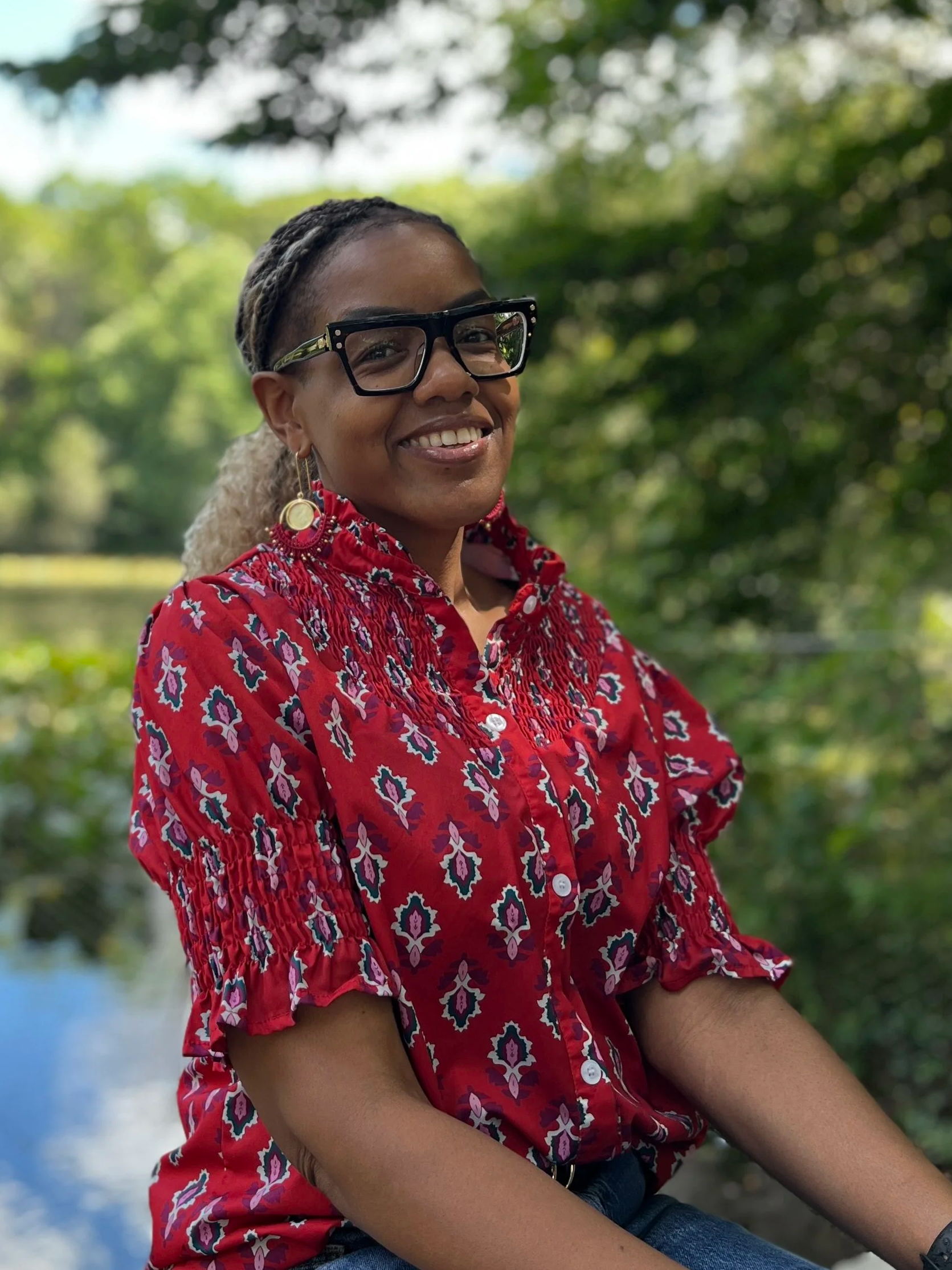 smiling woman with glasses and earrings sitting outdoors near water, wearing a red patterned blouse