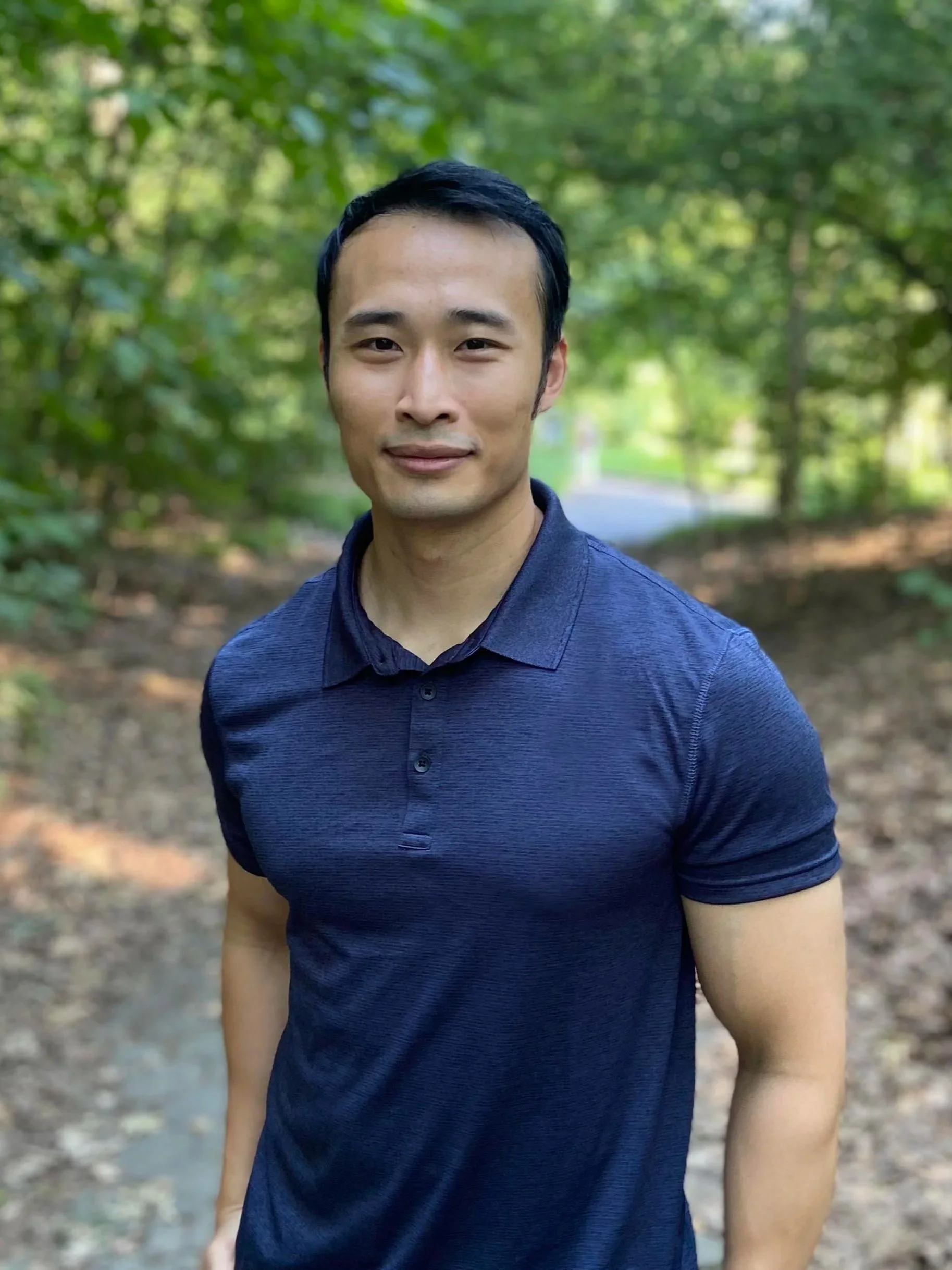 A man standing outdoors on a wooded trail, wearing a navy polo shirt, with trees and greenery in the background.