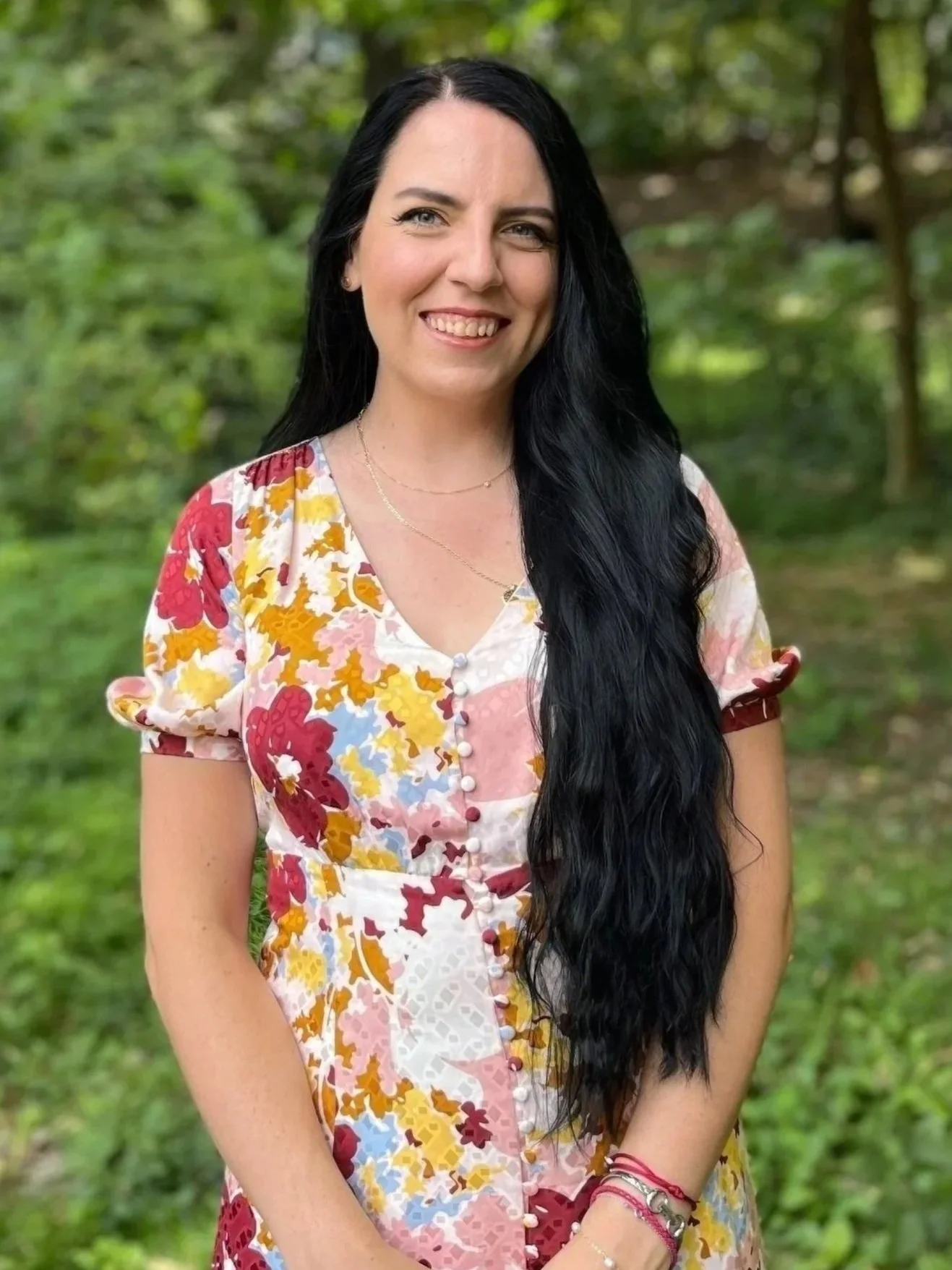 A woman with long black hair outdoors in a green, wooded area, smiling, wearing a colorful, floral dress with short puffed sleeves.