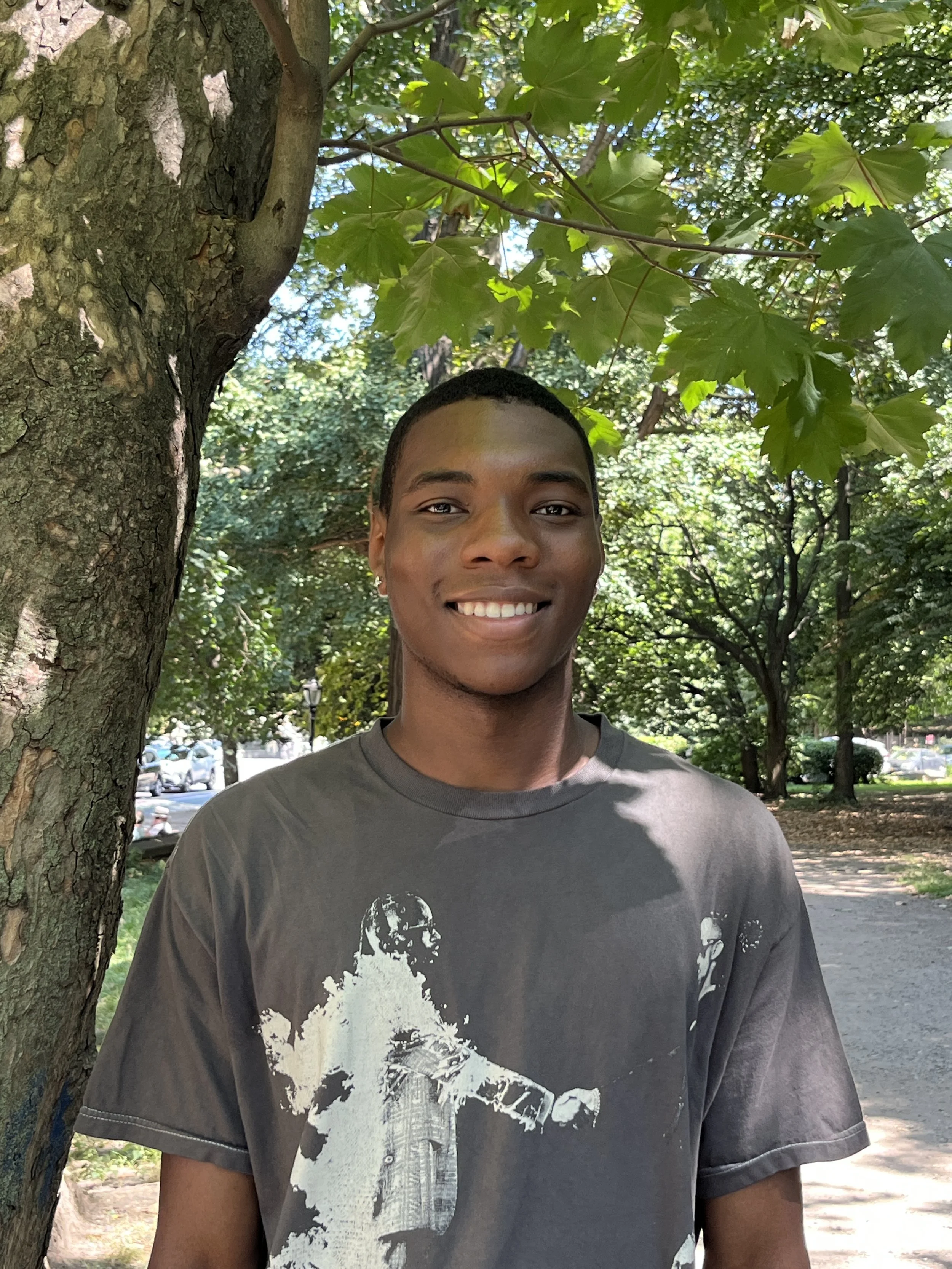 A young man smiling and standing outdoors next to a tree with green leaves.