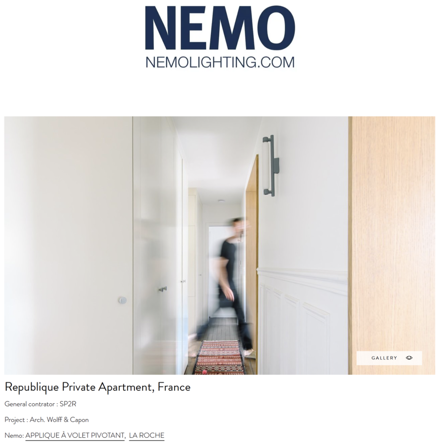 Une photo d'un couloir d'appartement avec un mur blanc et un sol en bois sombre. Un homme flou marche dans le couloir, passant devant une porte en bois. Il y a une lumière moderne fixée au mur à droite.