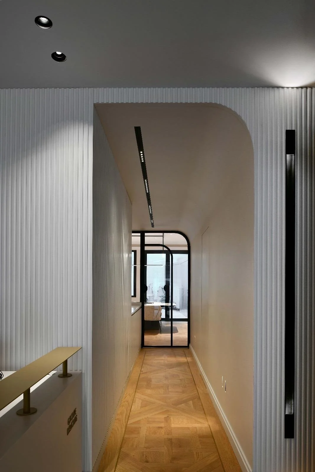 Un couloir moderne avec murs en plâtre blanc, sol en parquet de bois clair, plafond incurvé, éclairage encastré, et porte en verre noir à l'arrière.