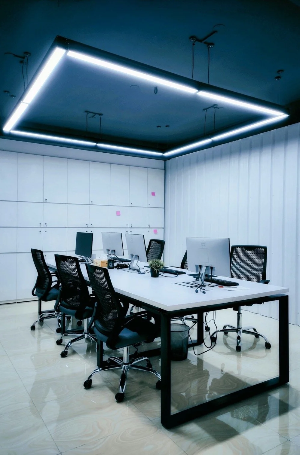 Salle de réunion moderne avec une table blanche, six chaises de bureau, plusieurs ordinateurs, un luminaire encastré au plafond, et un mur blanc avec des notes adhesives