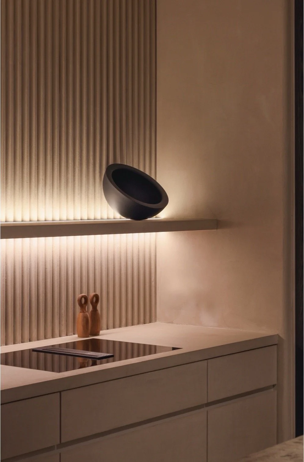 Une cuisine moderne avec un plan de travail en pierre beige, deux figurines en bois, une plaque de cuisson électrique et une décoration minimaliste avec un mur en bois ajouré et un objet noir arrondi posé en équilibre.