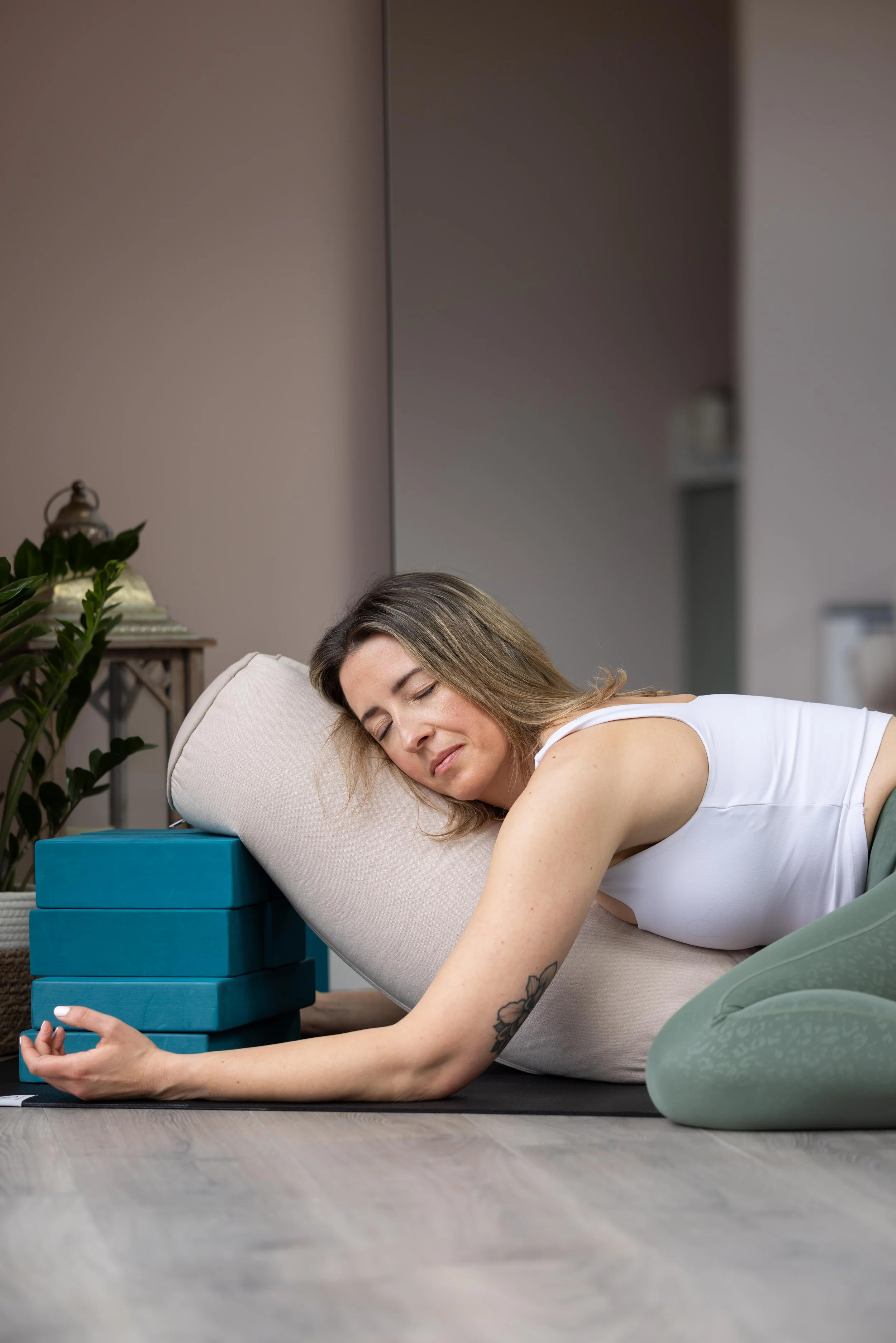A woman in a white tank top and green leggings is resting on a yoga bolster supported by blocks, relaxed with closed eyes, in a calming interior with plants and a lantern in the background.