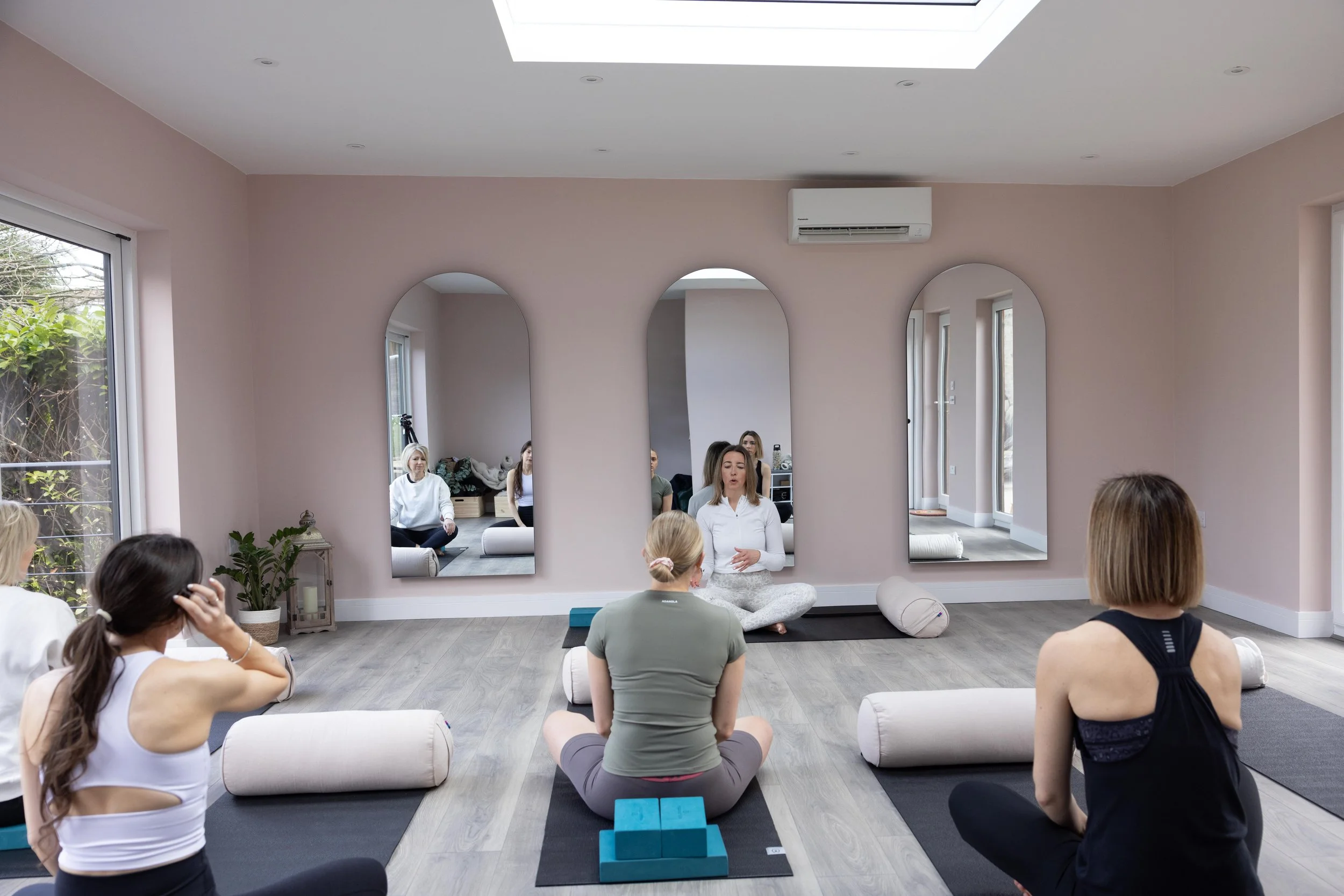 A group of people in a yoga or meditation class seated on mats in a studio with pink walls. The room has large mirrors, floor cushions, and natural light. A female instructor is leading the session.