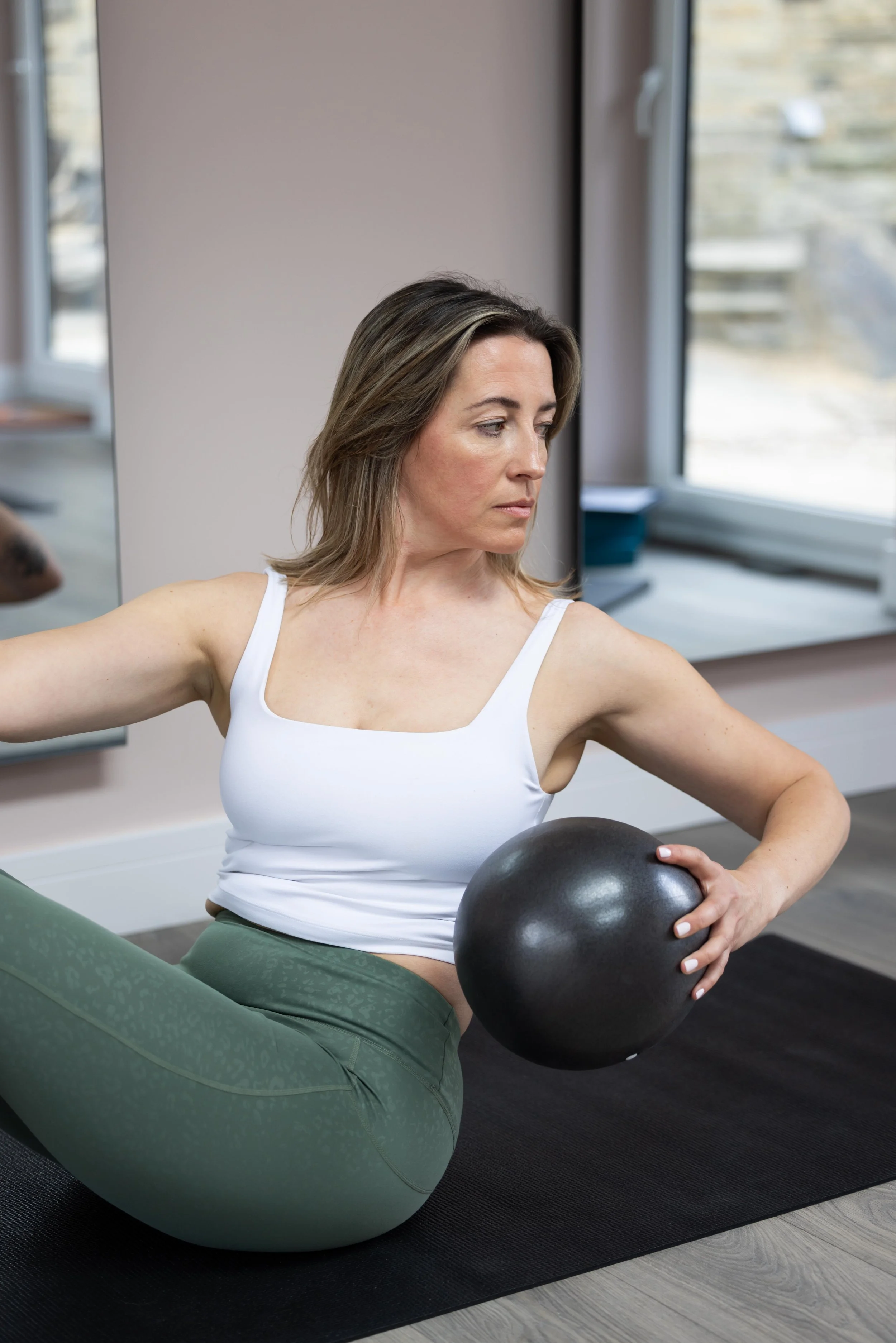 Woman in a white sports bra and green leggings exercising with a black fitness ball indoors.