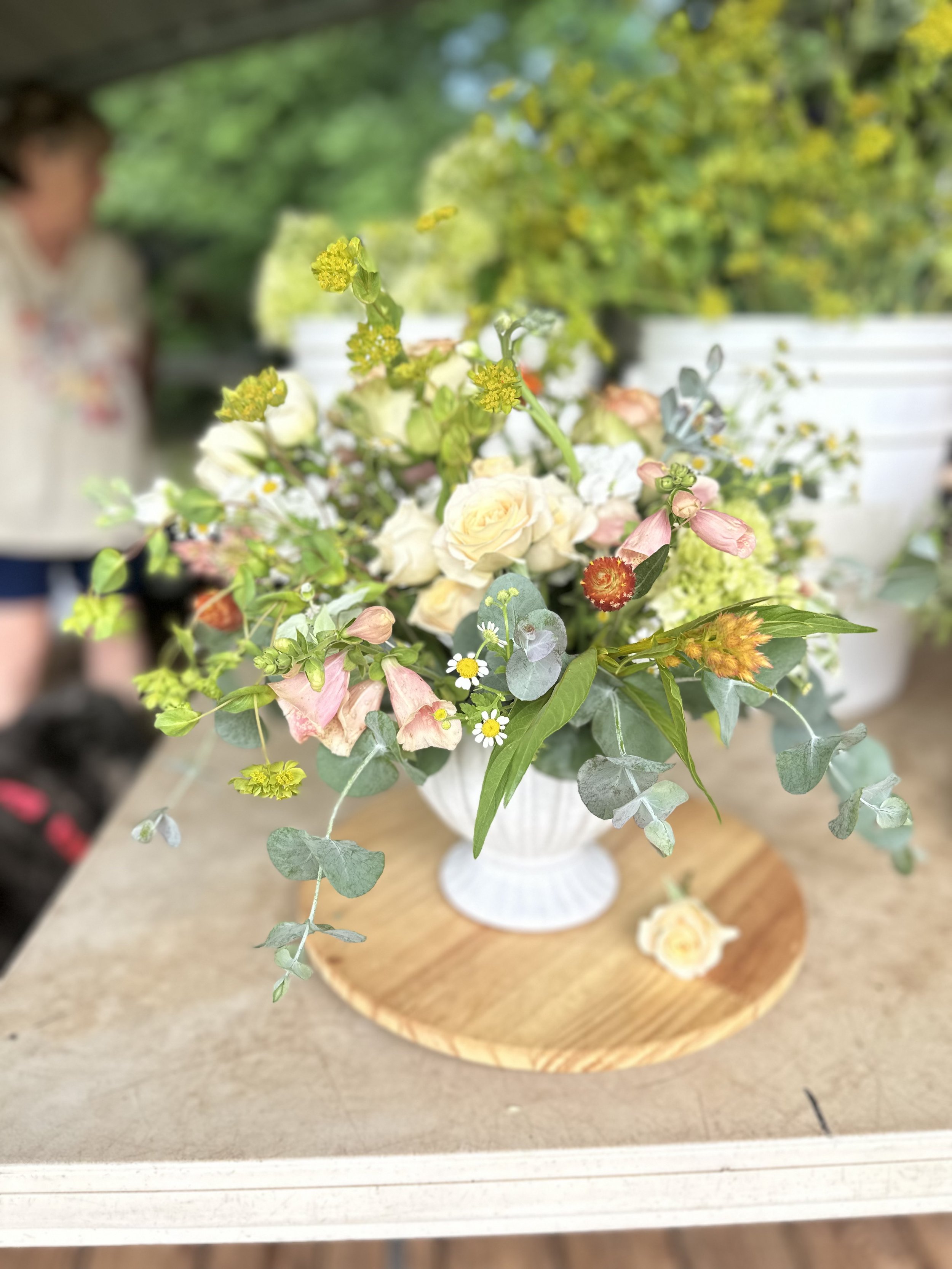 A floral arrangement in a white vase placed on a wooden base, featuring white roses, pink and orange flowers, small white daisy-like blooms, green foliage, and eucalyptus leaves. The arrangement is set on a table with a blurred background of greenery and a person.