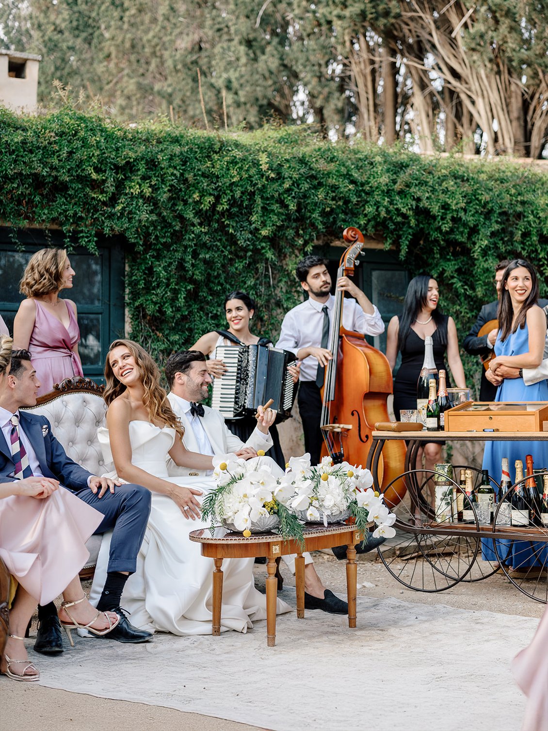 A group of people at a wedding reception, some sitting on a sofa and others standing, playing musical instruments, and celebrating outdoors with flowers and drinks.