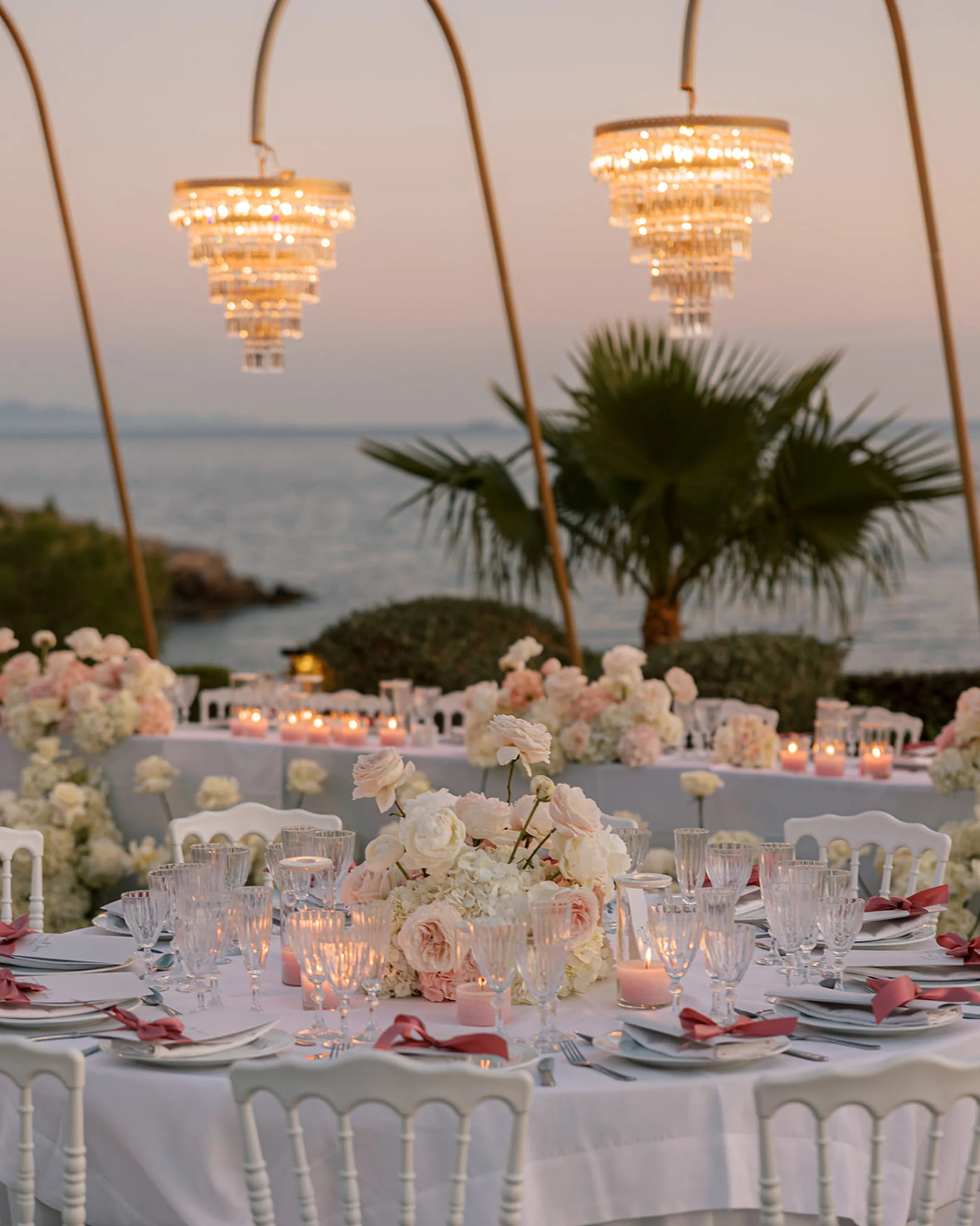 Elegant outdoor wedding reception table set with white tablecloth, glassware, floral centerpieces, and pink ribbons, overlooking the ocean with palm trees and chandeliers hanging overhead.