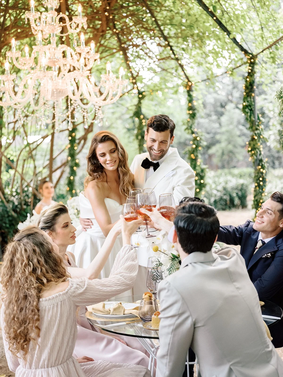 People celebrating at an outdoor wedding reception, clinking glasses of rosé wine under a chandelier.