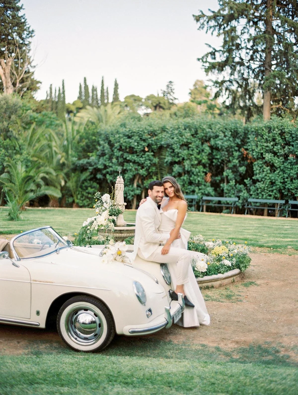 A couple in wedding attire sitting on the front of a vintage white convertible car decorated with white flowers, outdoors in a garden setting with lush green trees and bushes in the background.
