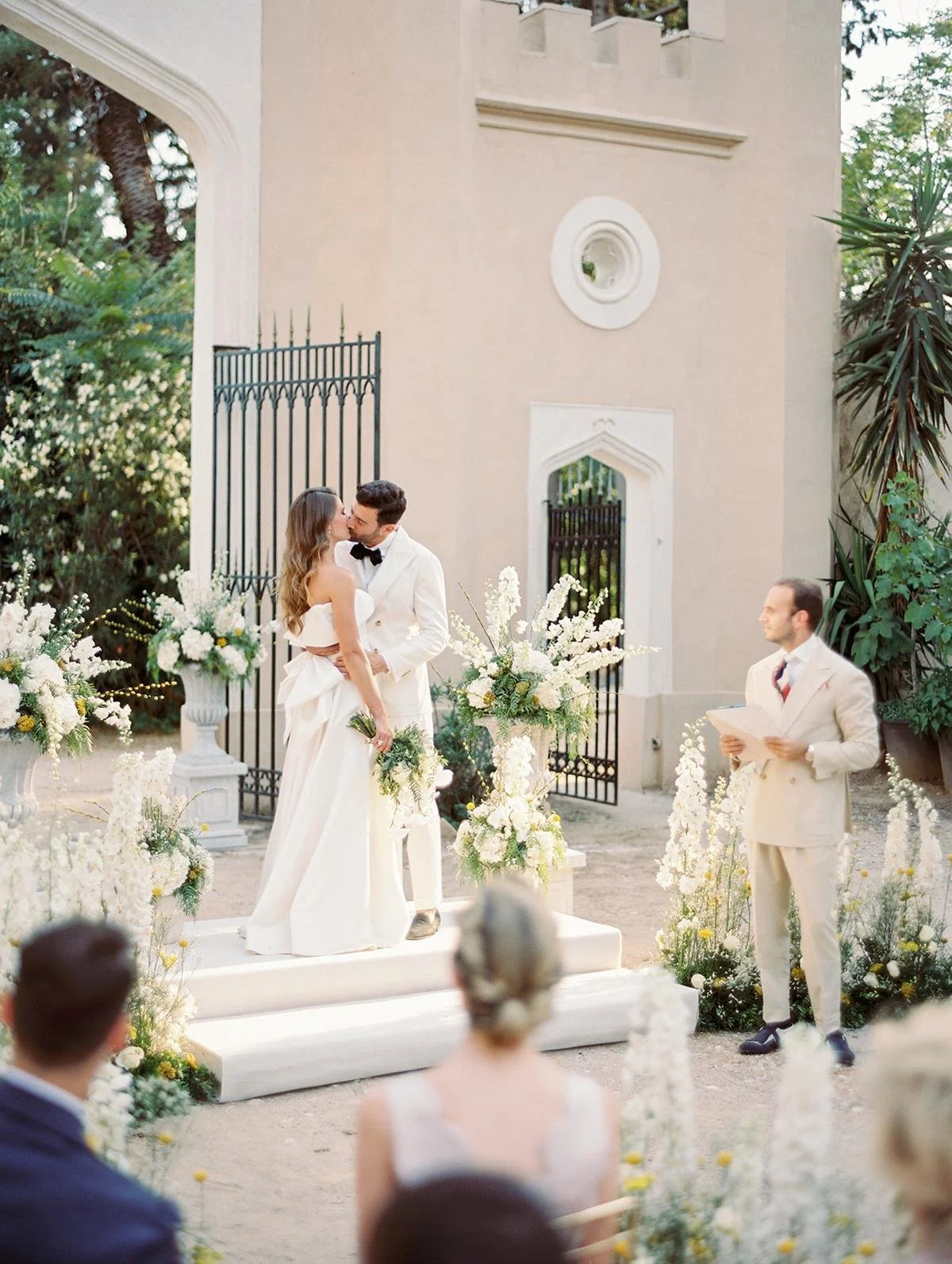 A wedding ceremony outdoors with a bride and groom kissing at the altar, surrounded by white flowers, with guests seated and a man officiating the service.