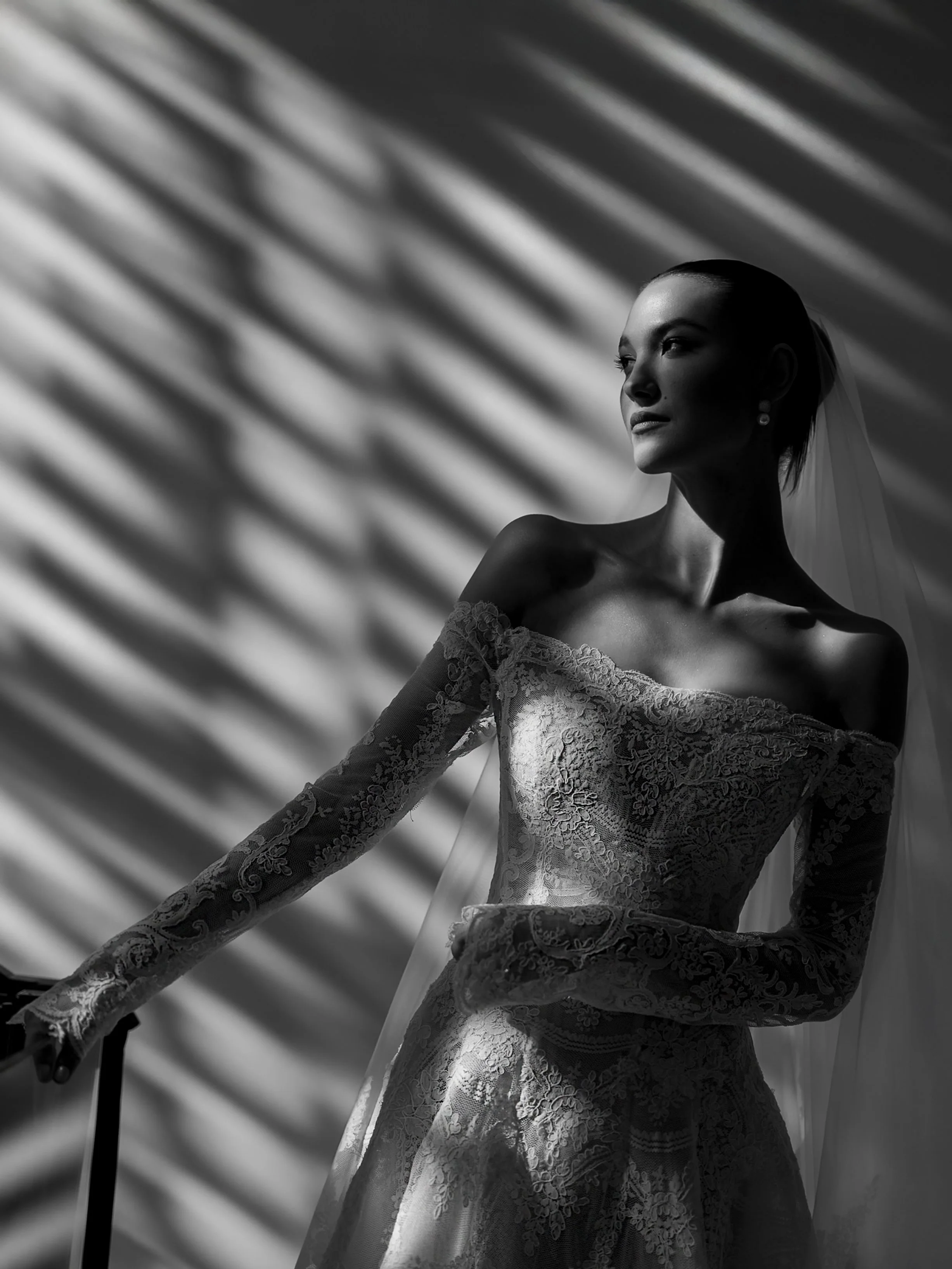 A woman in an off-shoulder lace wedding dress with long lace sleeves, standing in front of a window with patterned shadows, wearing a veil and pearl earrings.