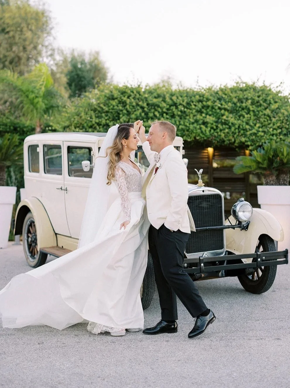 A bride and groom posing in front of a vintage white car outdoors, with lush greenery in the background, during their wedding celebration.