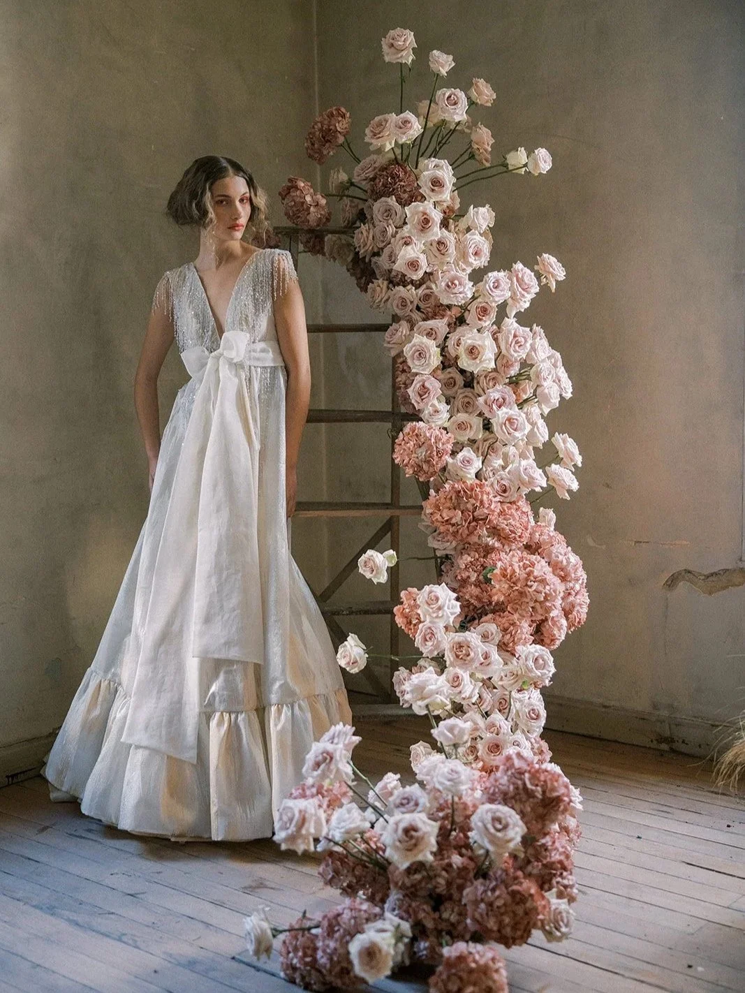 A young woman in a long, elegant white dress with lace and ribbon details standing next to a large floral display of pink and white roses and hydrangeas in an indoor setting with wooden flooring and plain walls.