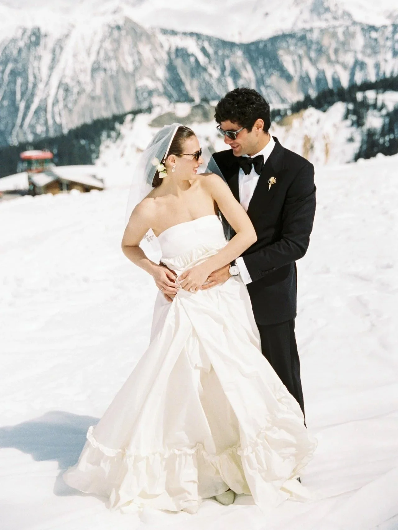 A bride and groom in wedding attire standing in the snow with snow-covered mountains in the background. The bride is wearing a strapless white wedding gown and veil, while the groom is dressed in a black tuxedo with sunglasses. They are holding hands