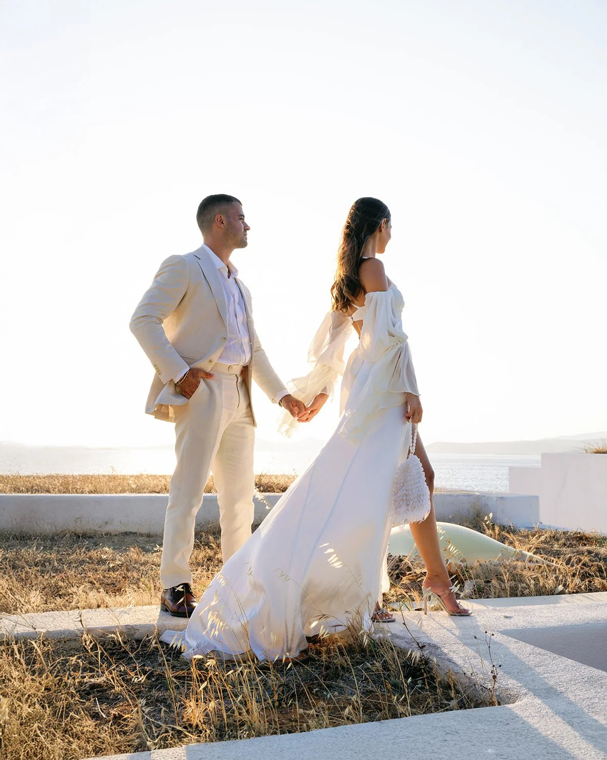 A couple dressed in wedding attire holding hands outdoors during sunset by the water.