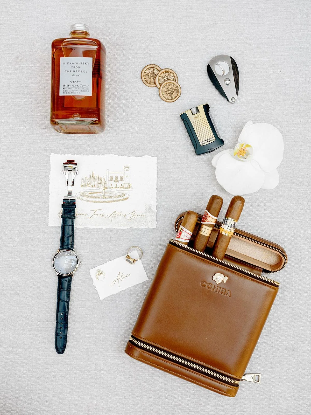 Collection of luxury items including a bottle of Nikka whisky, three gold coins, a silver and black bottle opener, a lighter, a white orchid, three cigars in a brown leather cigar case, a wristwatch with a leather strap, a handwritten note with "Alex