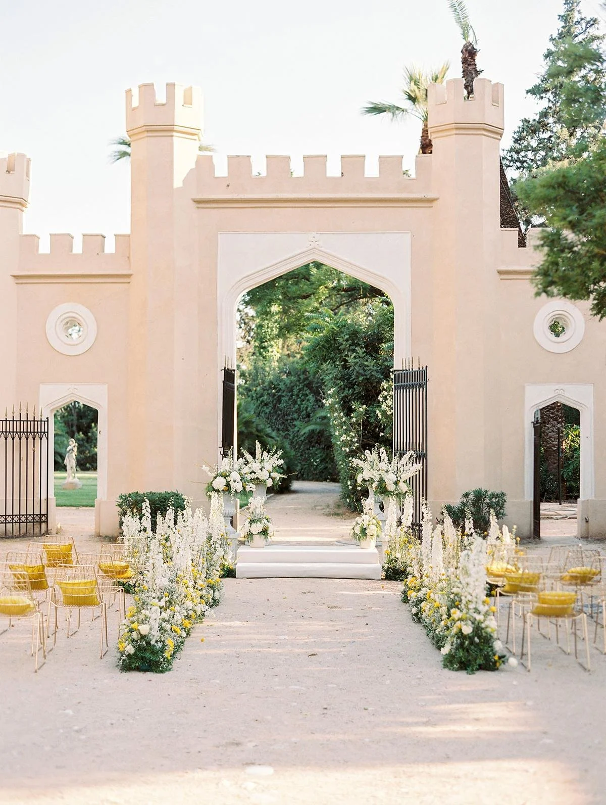 An outdoor wedding setup with an arched entrance, floral arrangements, and rows of transparent yellow chairs on either side of an aisle.