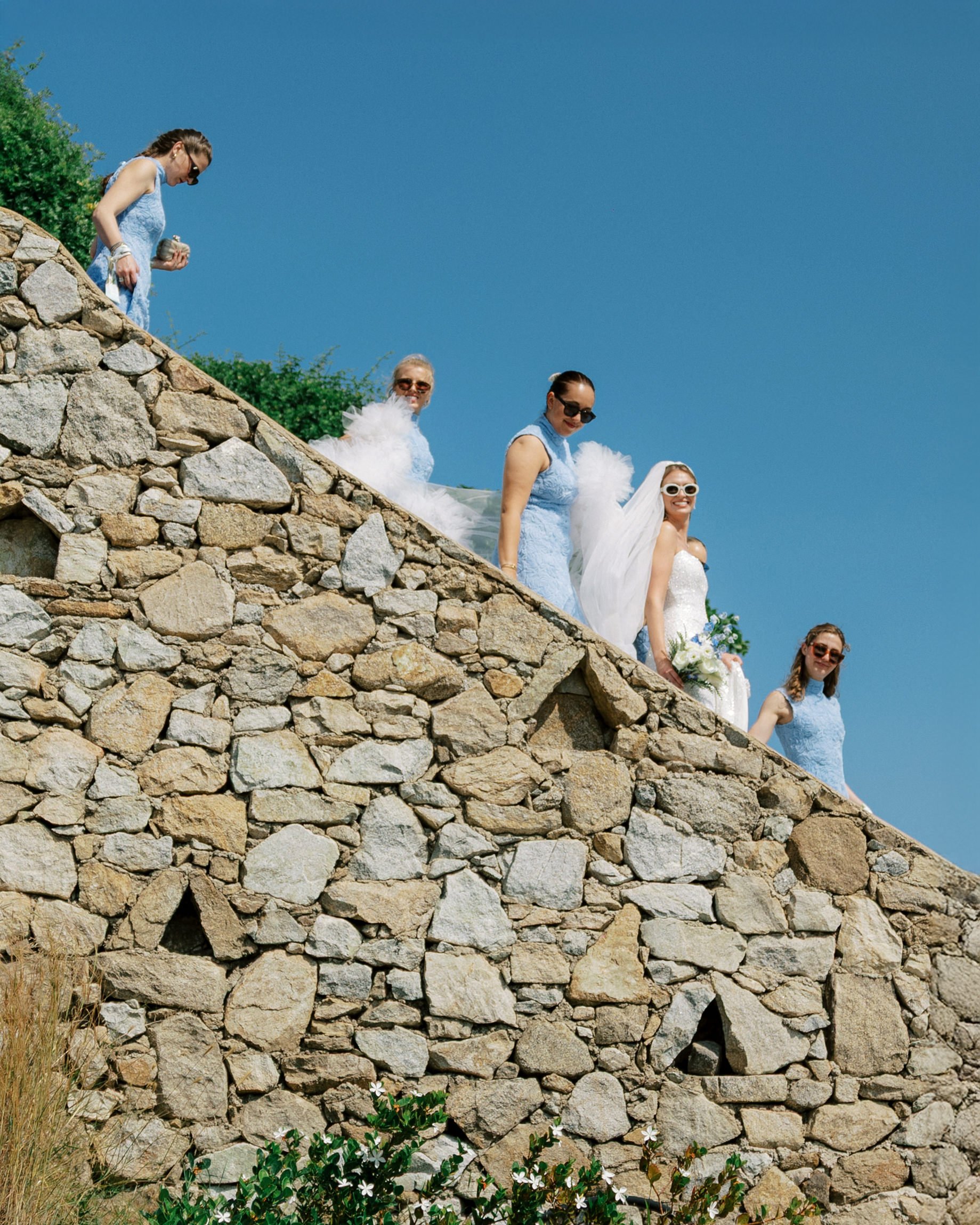 A wedding party walking down a stone staircase outdoors under a clear blue sky. The bride, wearing a white wedding dress and sunglasses, is holding a bouquet. Bridesmaids in blue dresses and sunglasses are accompanying her.