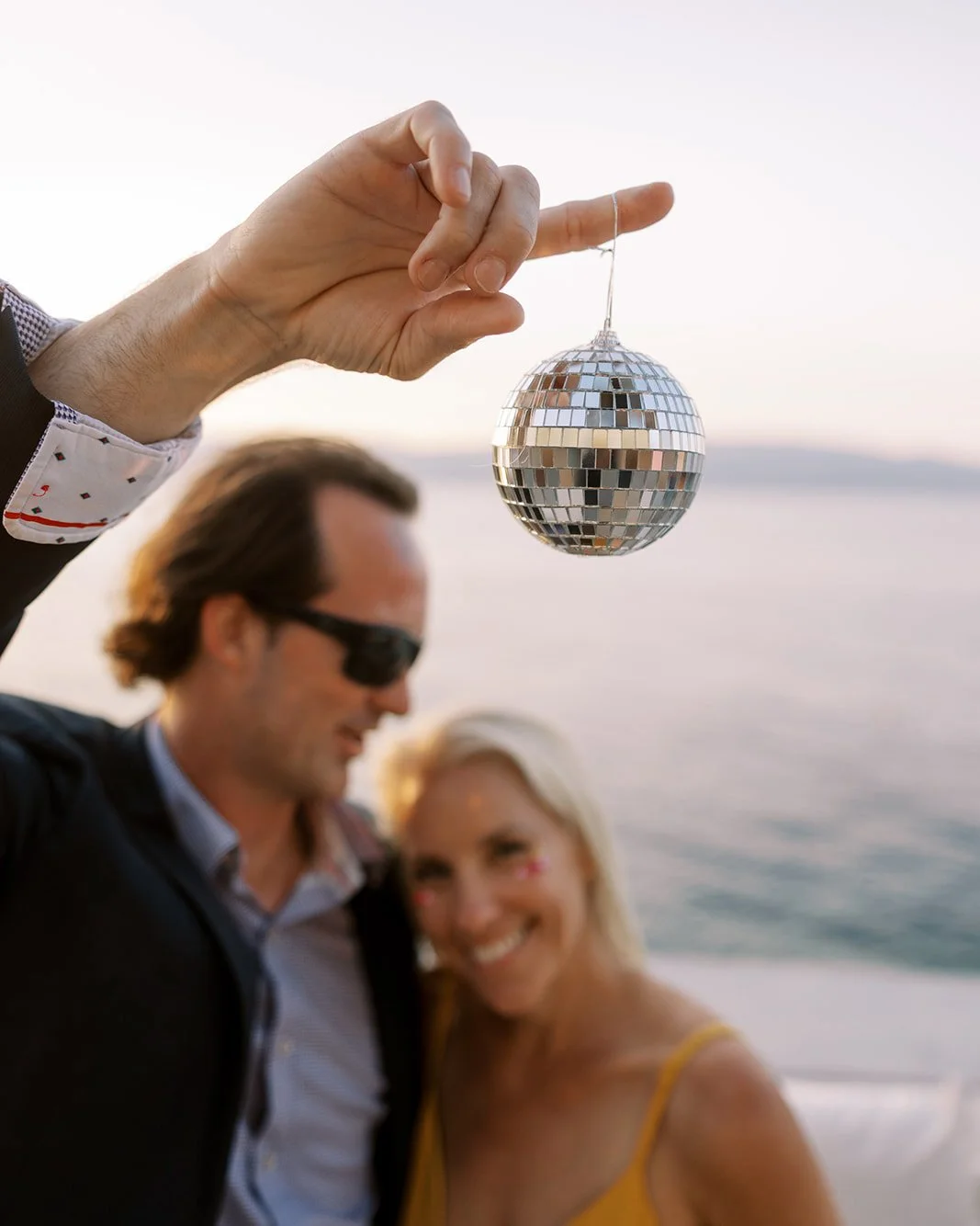 Person holding a small disco ball while a couple smiles in the background near the water during sunset.