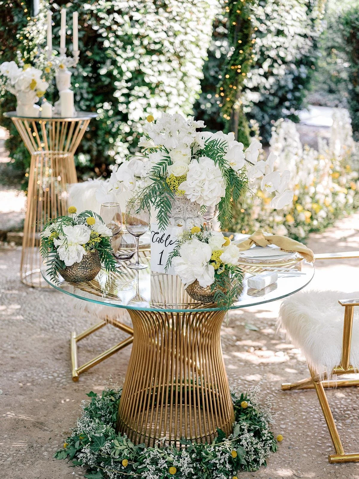 Elegant outdoor table setting with white floral arrangements, gold accents, glassware, and greenery, decorated for a wedding or special event.