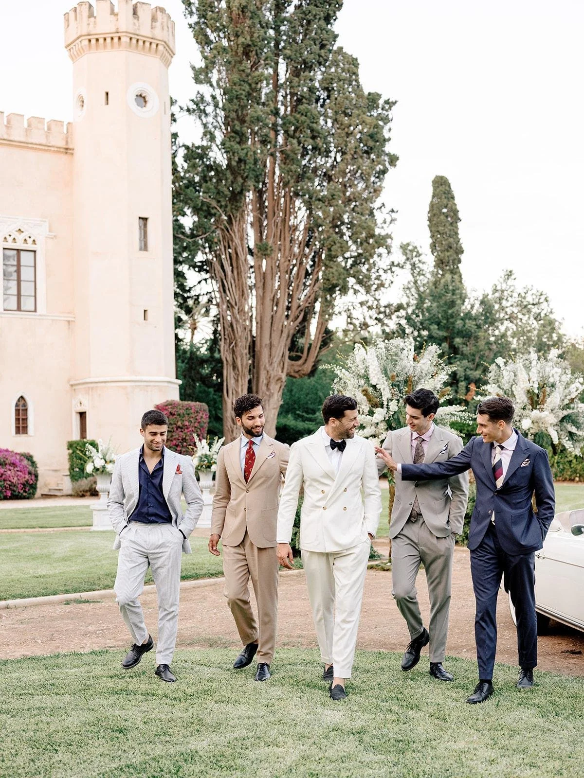 Group of six men dressed in suits walking outdoors in front of a mansion and large trees.