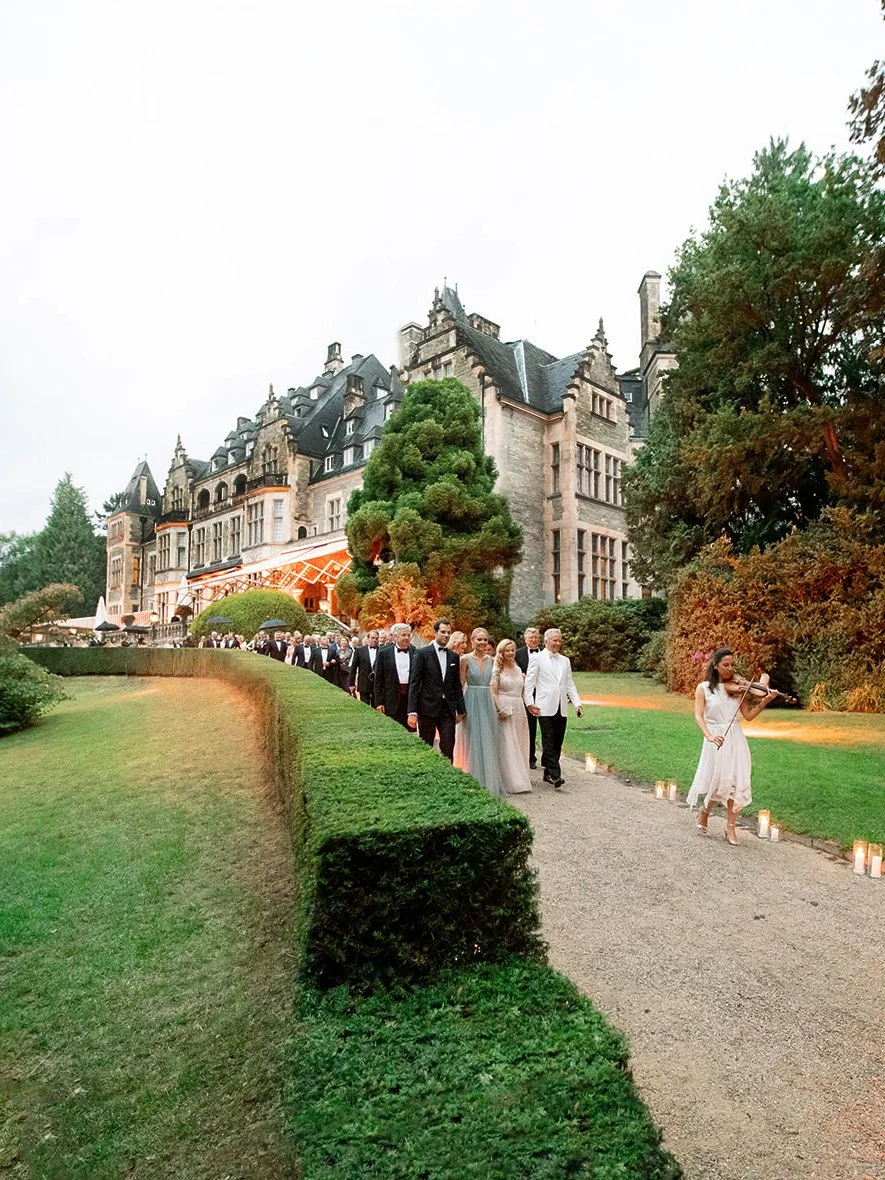 A group of people dressed in formal attire walking along a gravel path outside a large, historic castle-like building. A woman is playing a violin on the right side of the path, and small candles line the path.