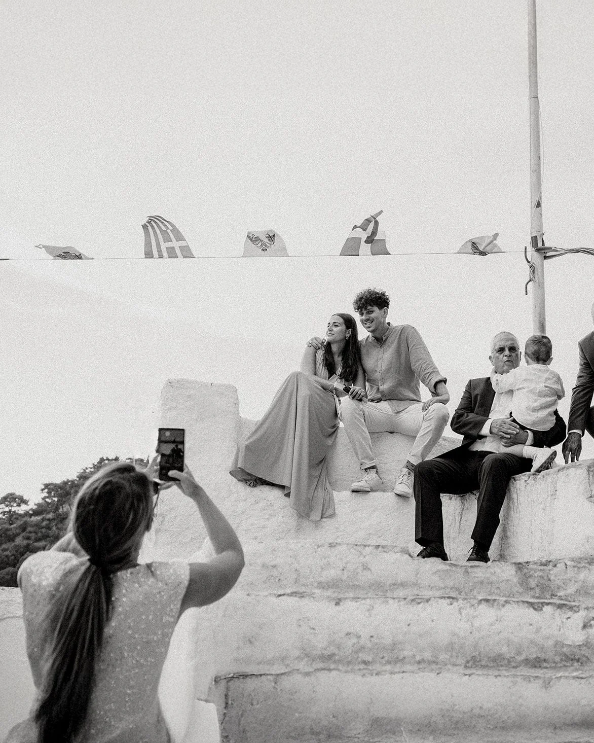 A woman takes a photo of a couple and an older man with a child sitting on his lap, all seated on stone steps outdoors during a sunny day.