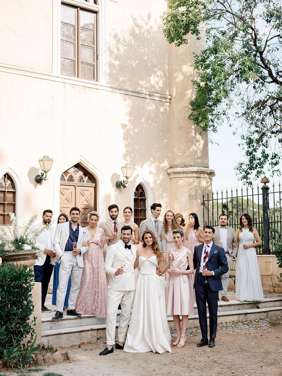 A group of people dressed in formal attire standing on steps outside a historic building celebrating a wedding or special event, with some holding glasses of wine.