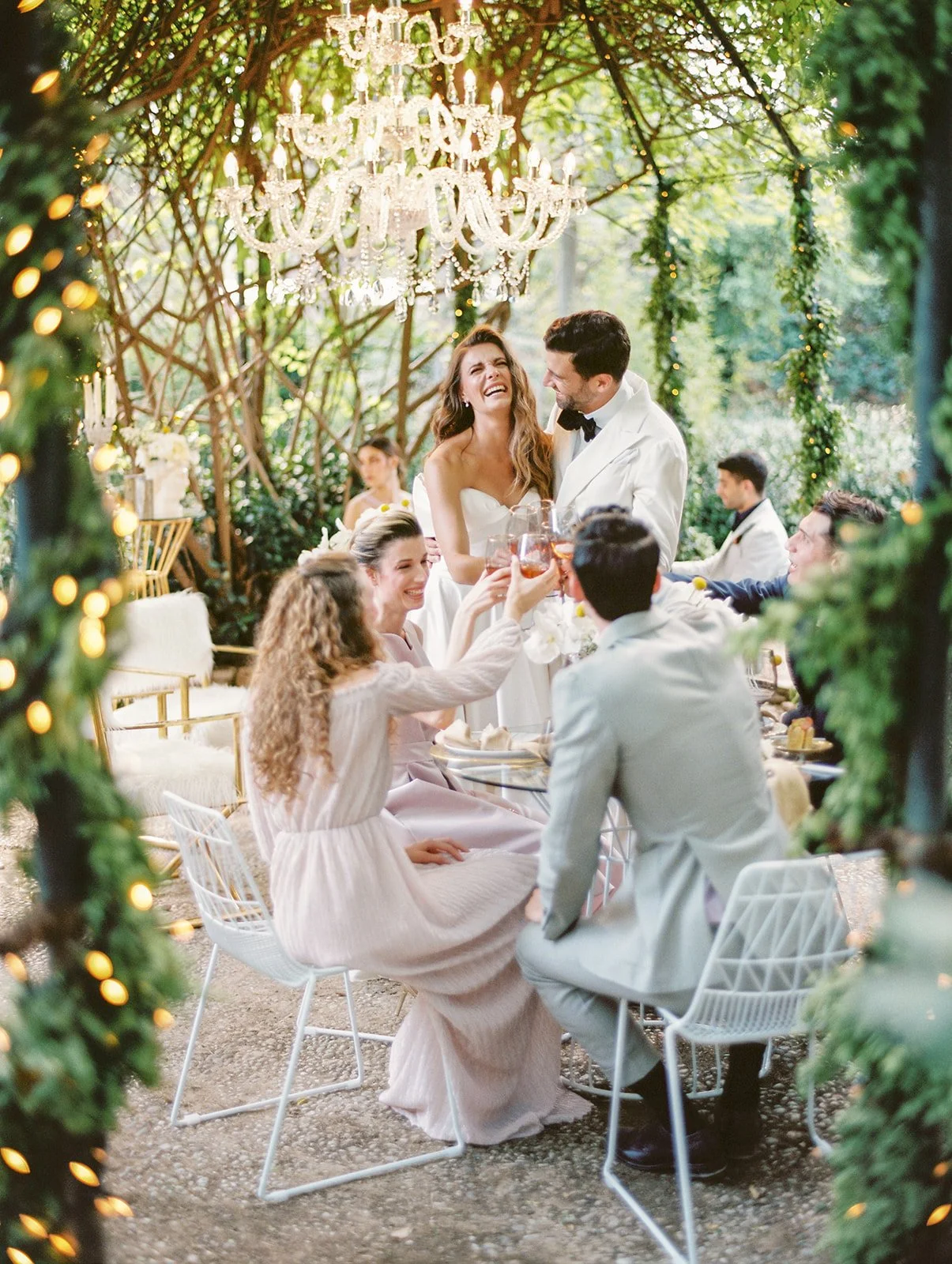 A wedding celebration taking place outdoors under a chandelier and surrounded by greenery, with a bride and groom toasting with friends.