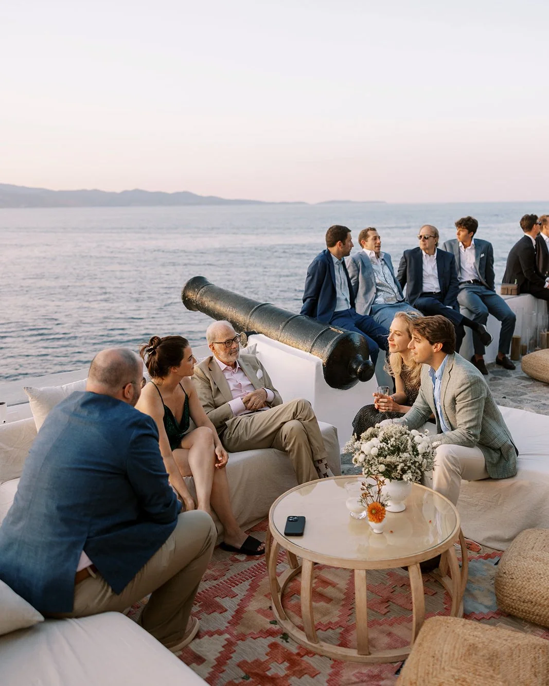 People gathered on a boat deck enjoying a sunset, with a cannon and waters in the background, engaging in conversation and holding drinks.