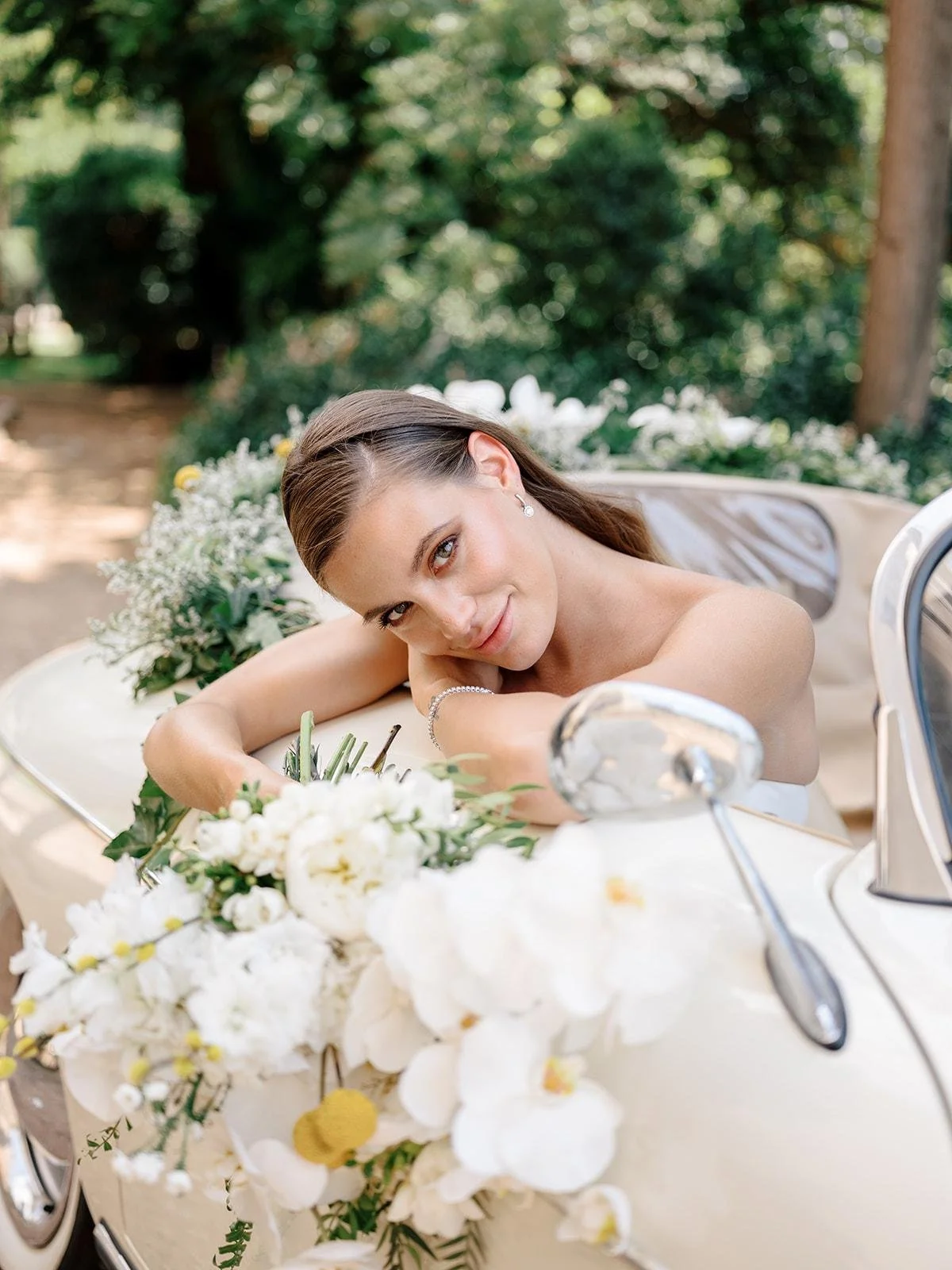 A woman lying in a vintage car decorated with white flowers, outdoors with green trees in the background.